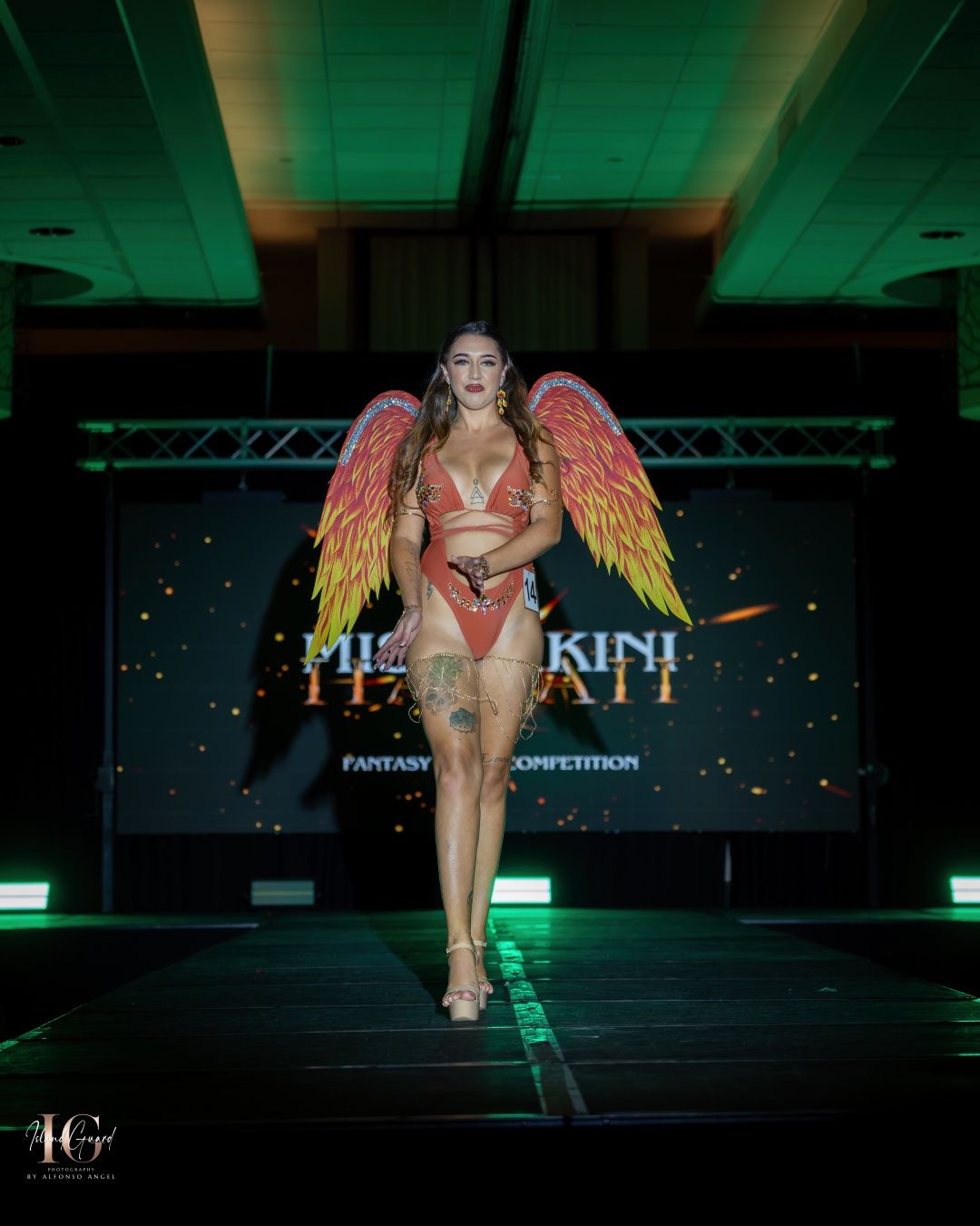 A model walks the runway wearing orange lingerie and dramatic golden wings in a fashion show with green lighting.