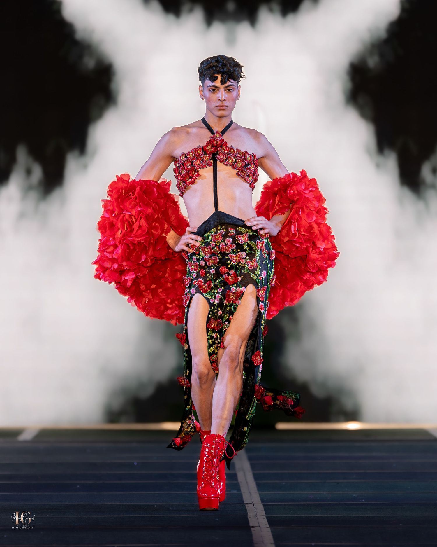 Dramatic fashion runway moment featuring red feathered accessories, floral print outfit and red boots amid theatrical fog effects.