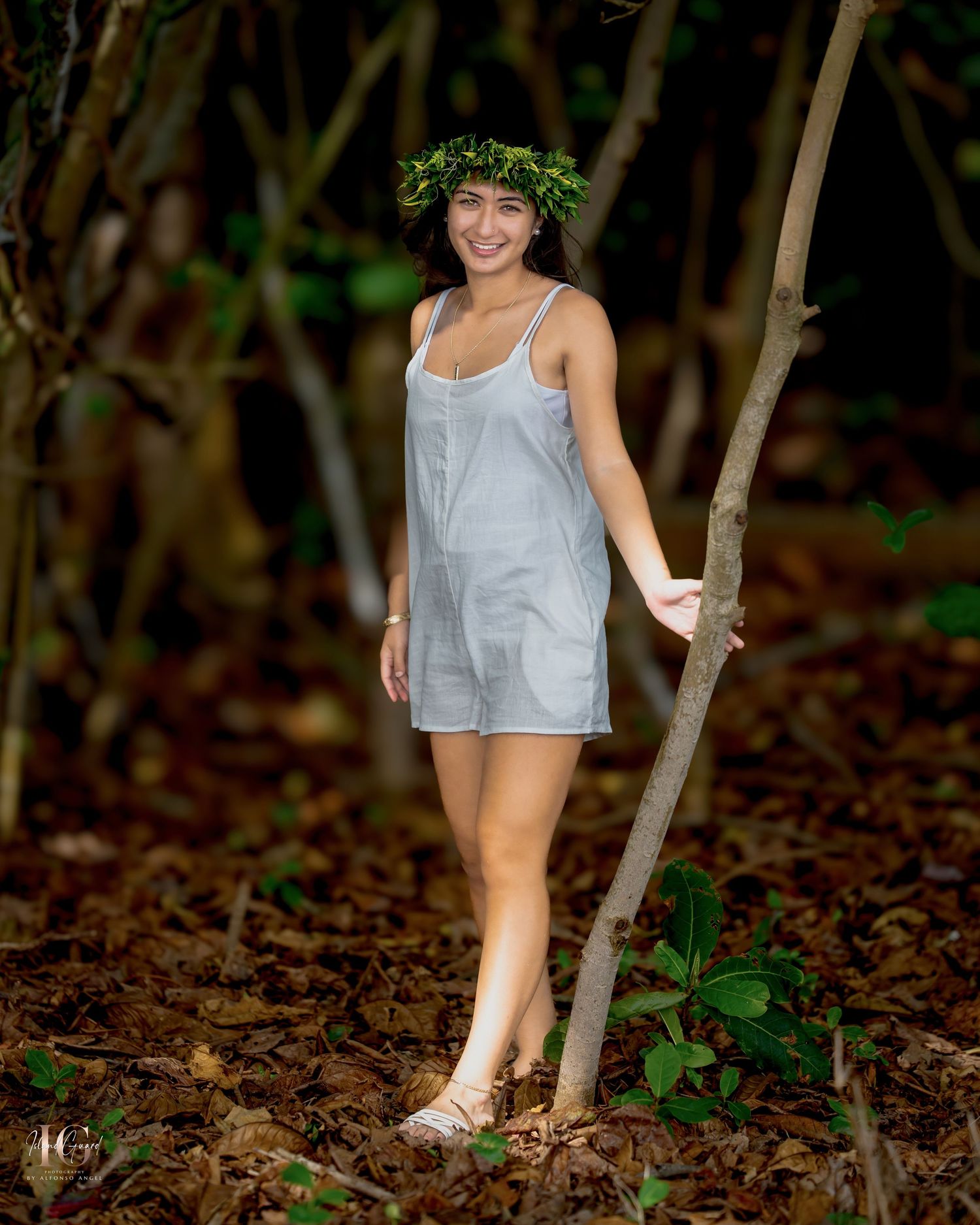 A person in light colored romper walks through autumn leaves scattered on forest ground holding tree branches.