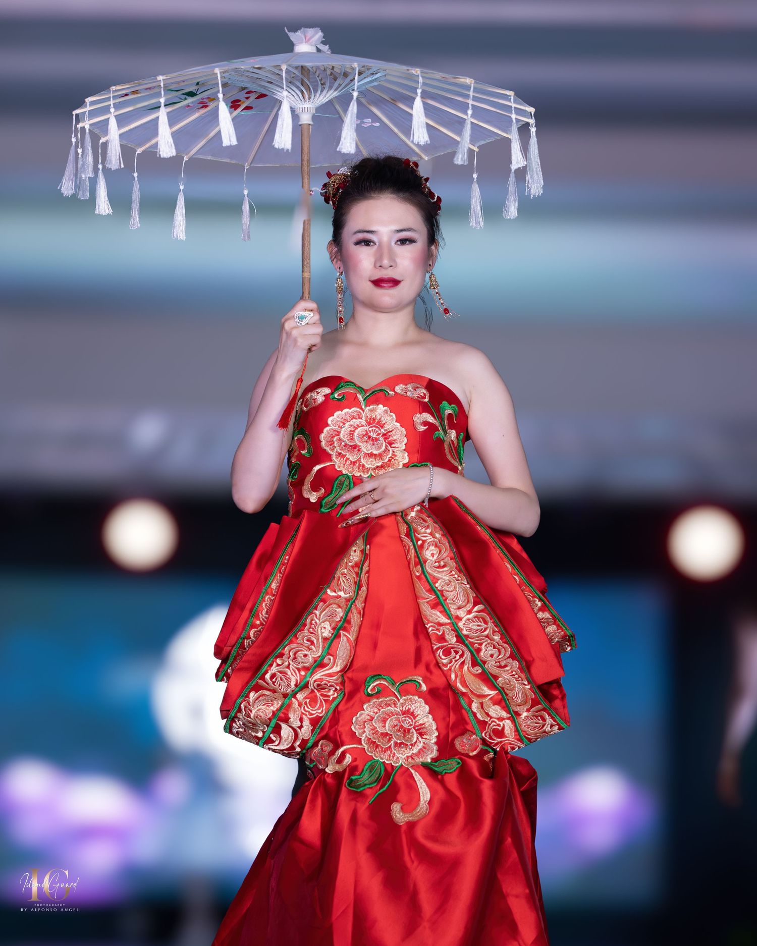 Model in red floral gown holds decorative white parasol on fashion runway with blue backdrop.
