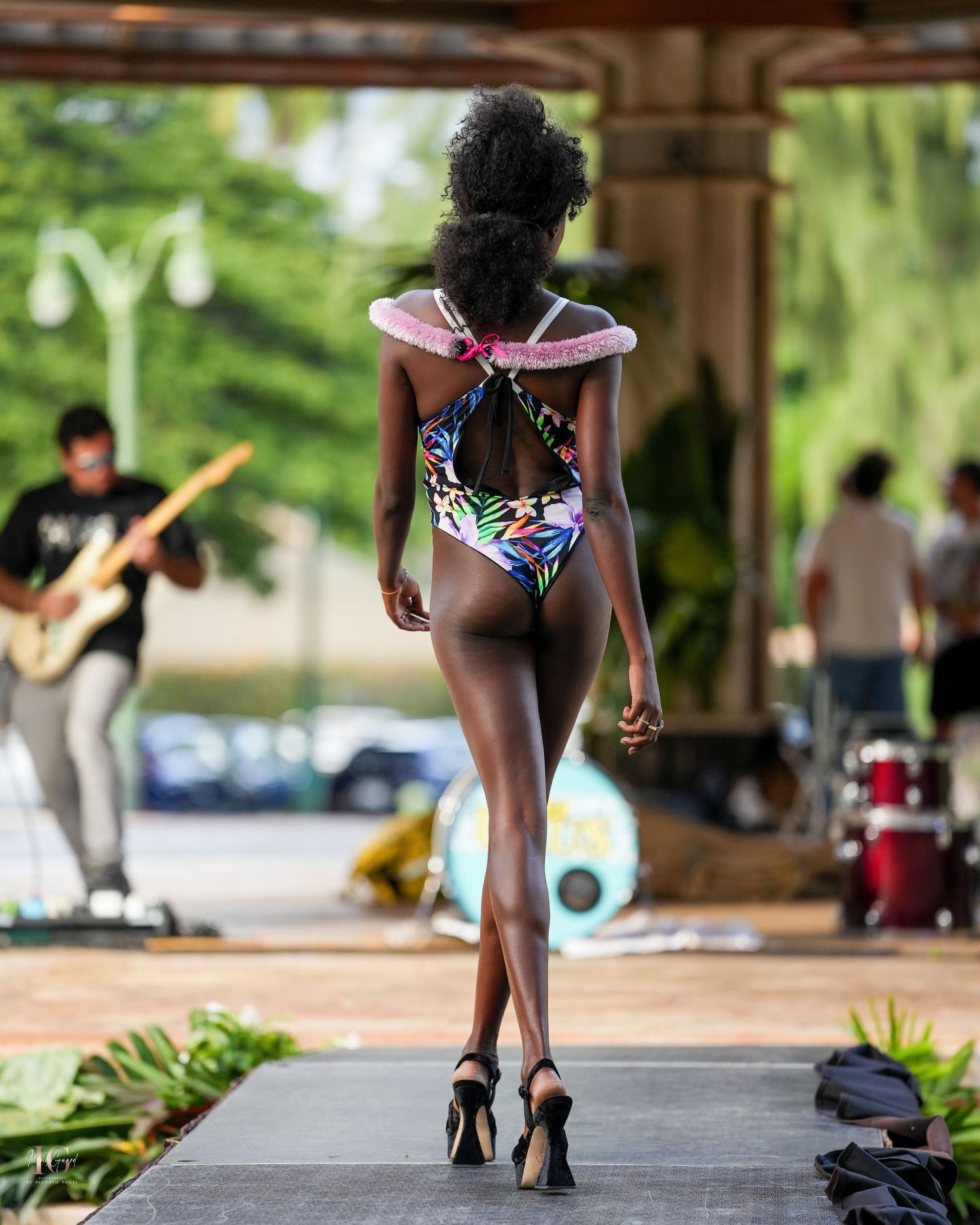 Model walks runway in floral swimwear and heels while musicians perform in background at outdoor fashion show.