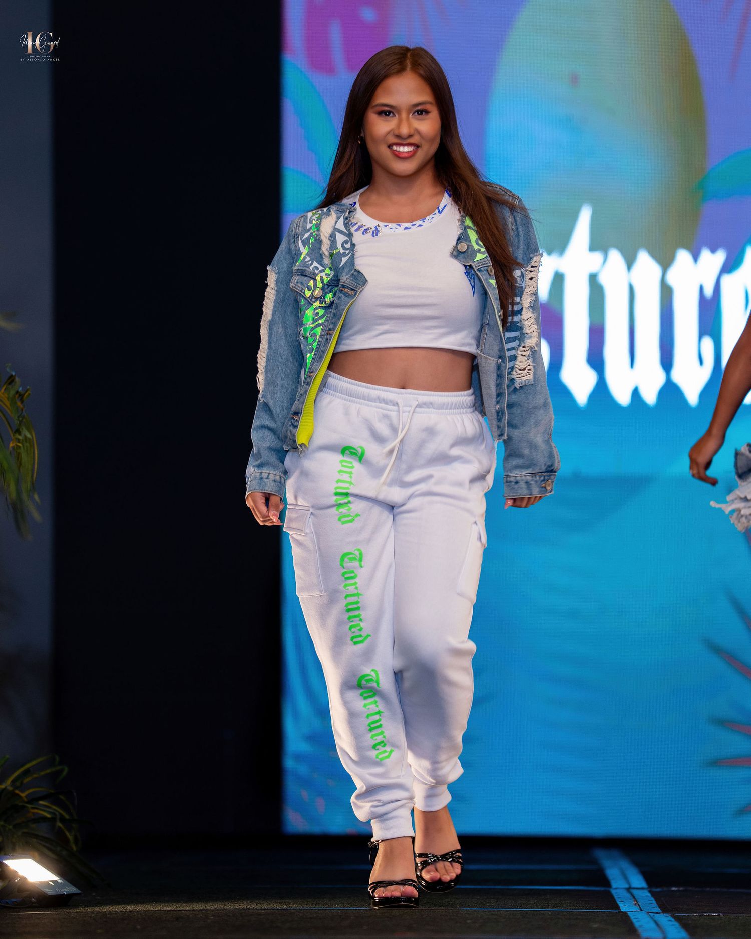 Model walks runway wearing white crop top, denim jacket with embellishments, and white sweatpants at fashion show.