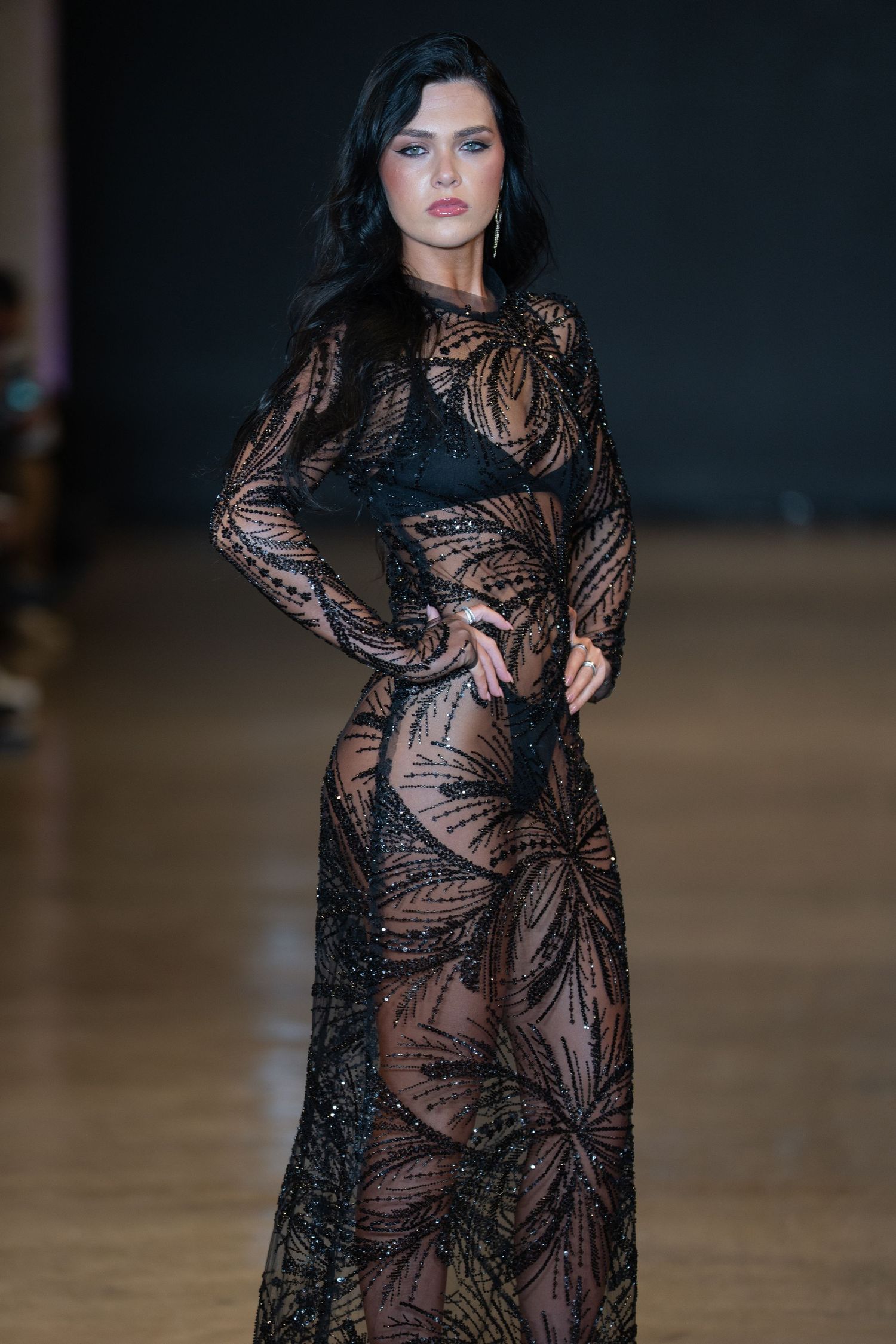 Fashion model walks runway in sheer black lace evening gown with intricate floral pattern against dark backdrop.