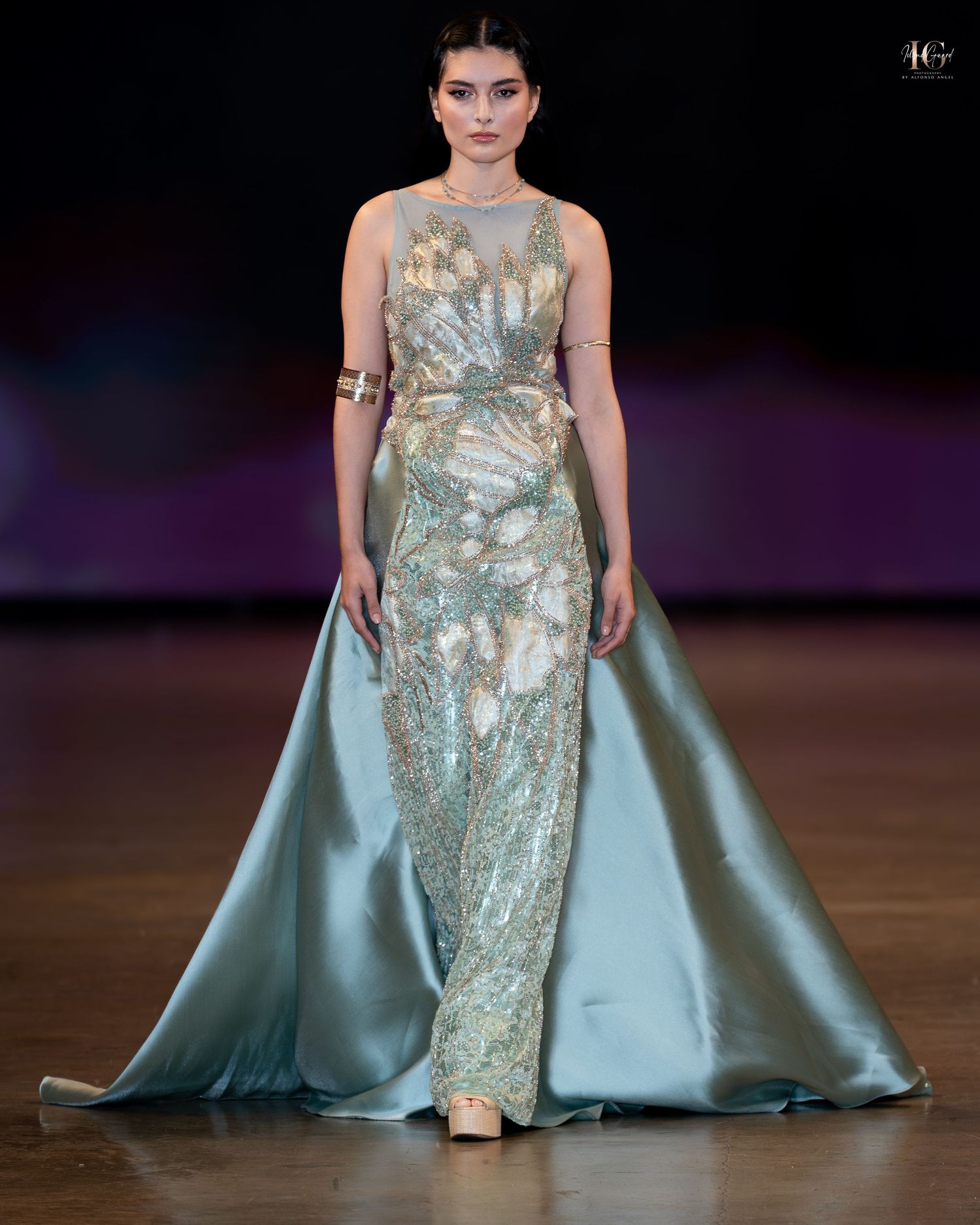 A model walks the runway in a shimmering silver and mint evening gown with dramatic cape detail at a fashion show.