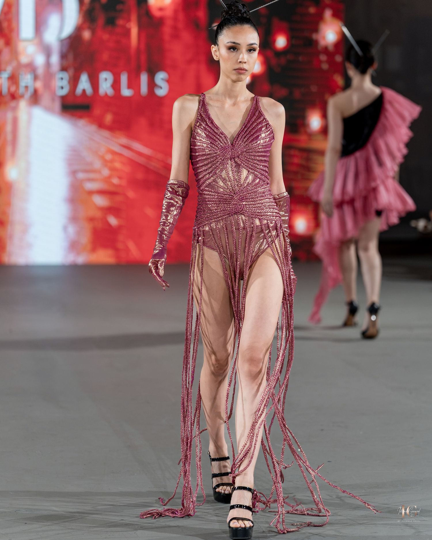 Model walks runway in sheer burgundy gown with dramatic fringe details at fashion show.