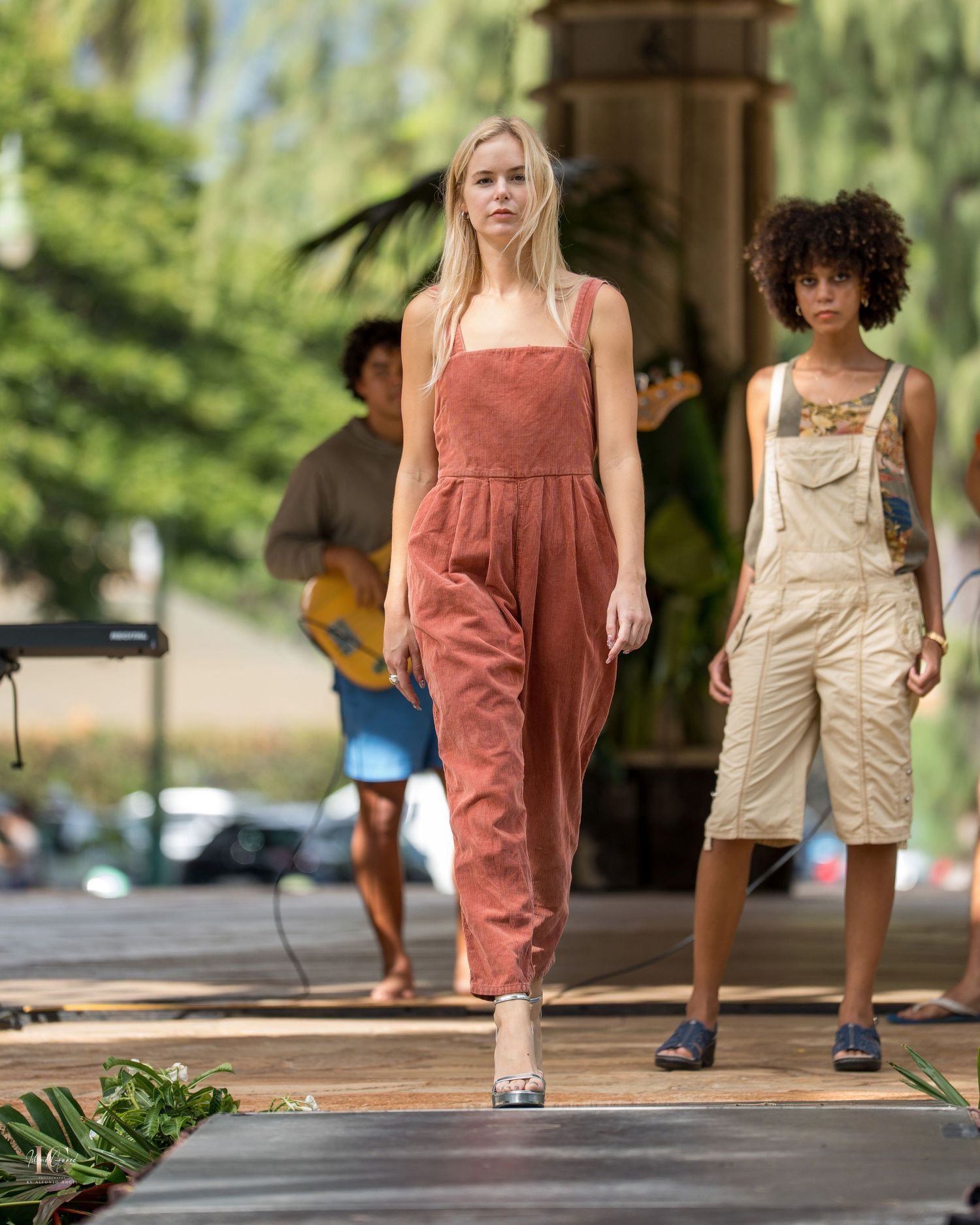 Models walk the runway wearing earth-toned summer dresses and casual wear during an outdoor fashion show.