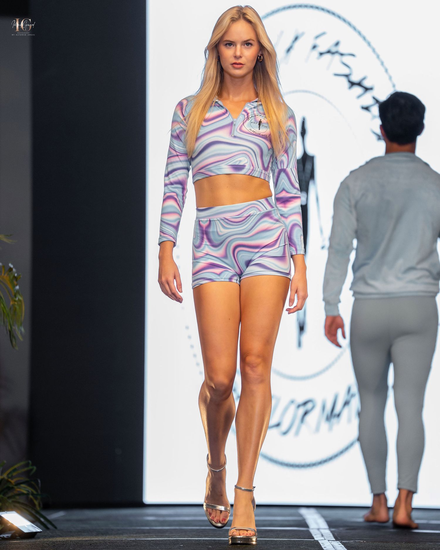 Model walks runway in purple and blue swirl print crop top and shorts set during fashion show.