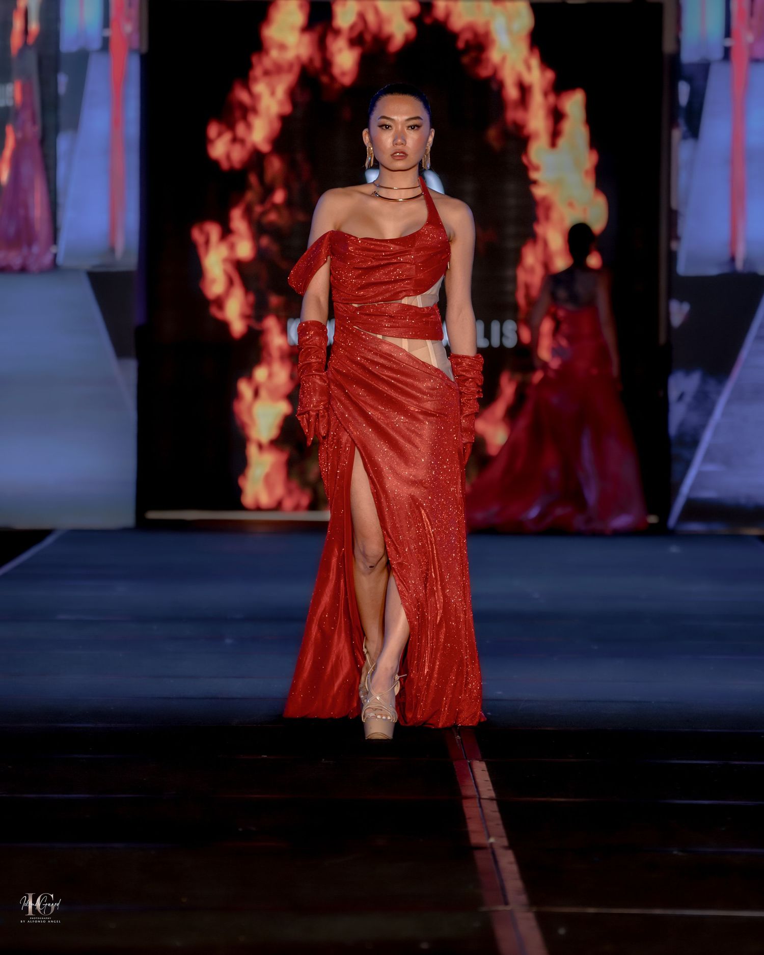 Model walks runway in sparkly red evening gown with thigh-high slit and matching gloves on illuminated catwalk stage.