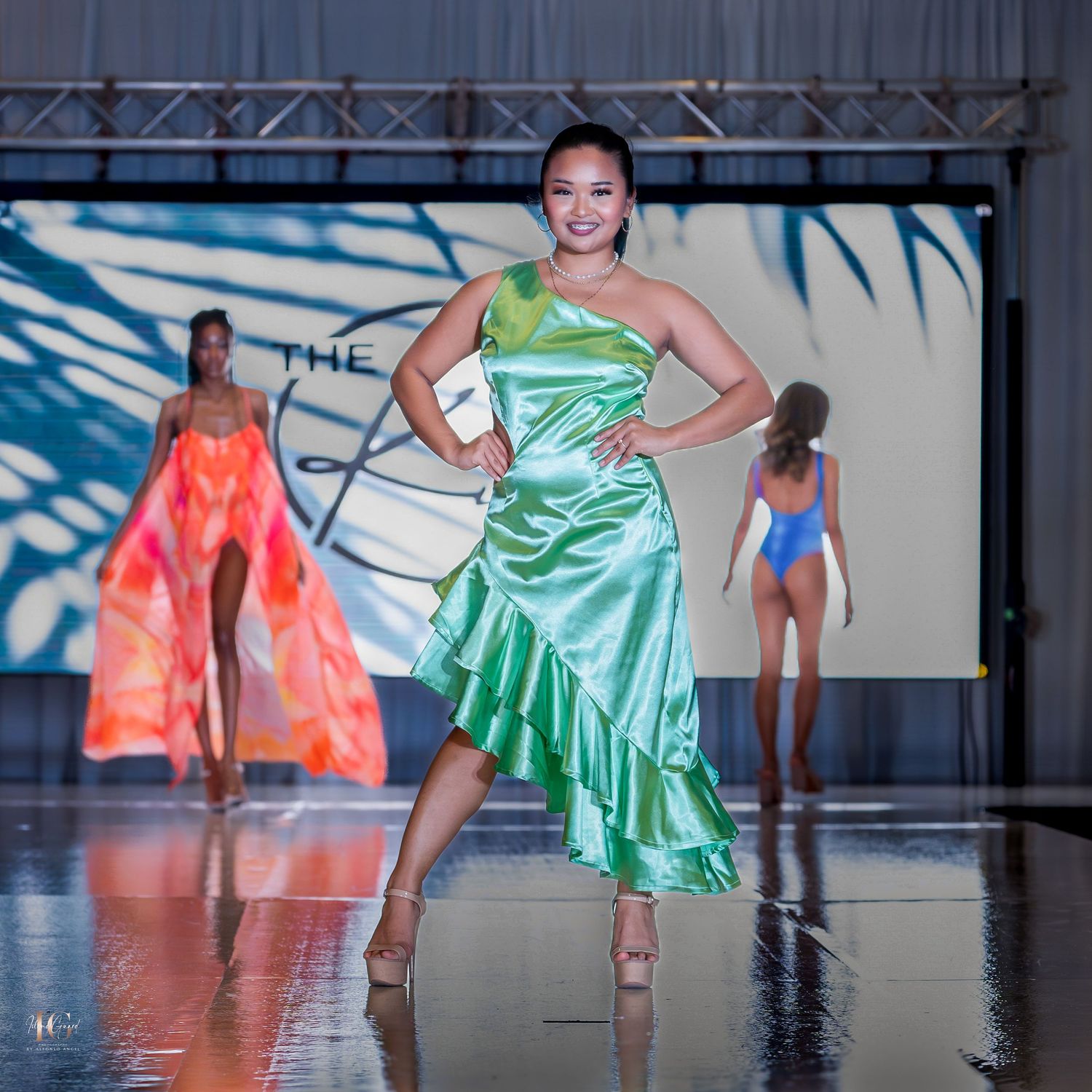 Models present metallic green swimwear and dresses on a reflective runway with dramatic lighting.