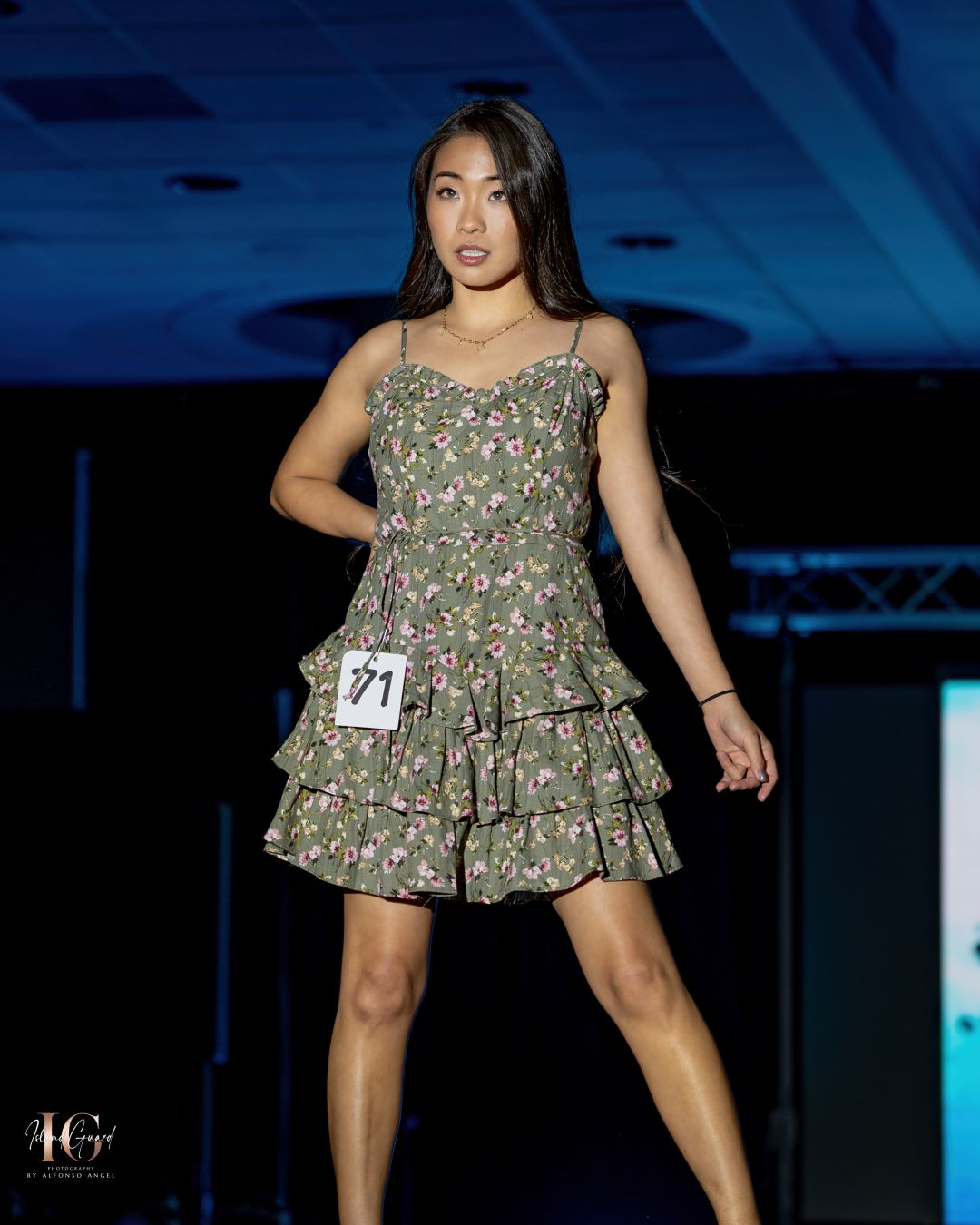 Model wearing a green floral tiered mini dress performs on runway with dark blue lighting at fashion show.