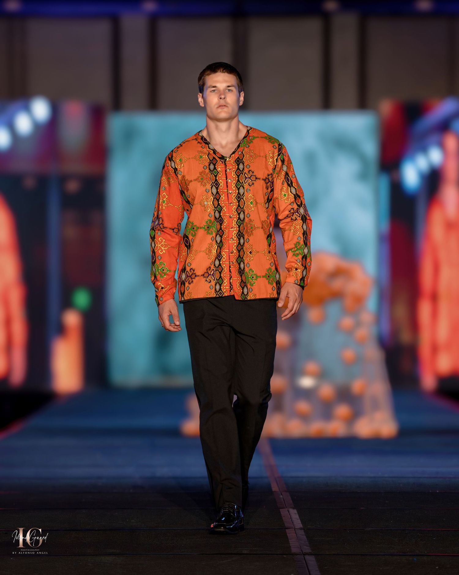 Model showcasing orange snakeskin-print jacket with black pants on dark catwalk.