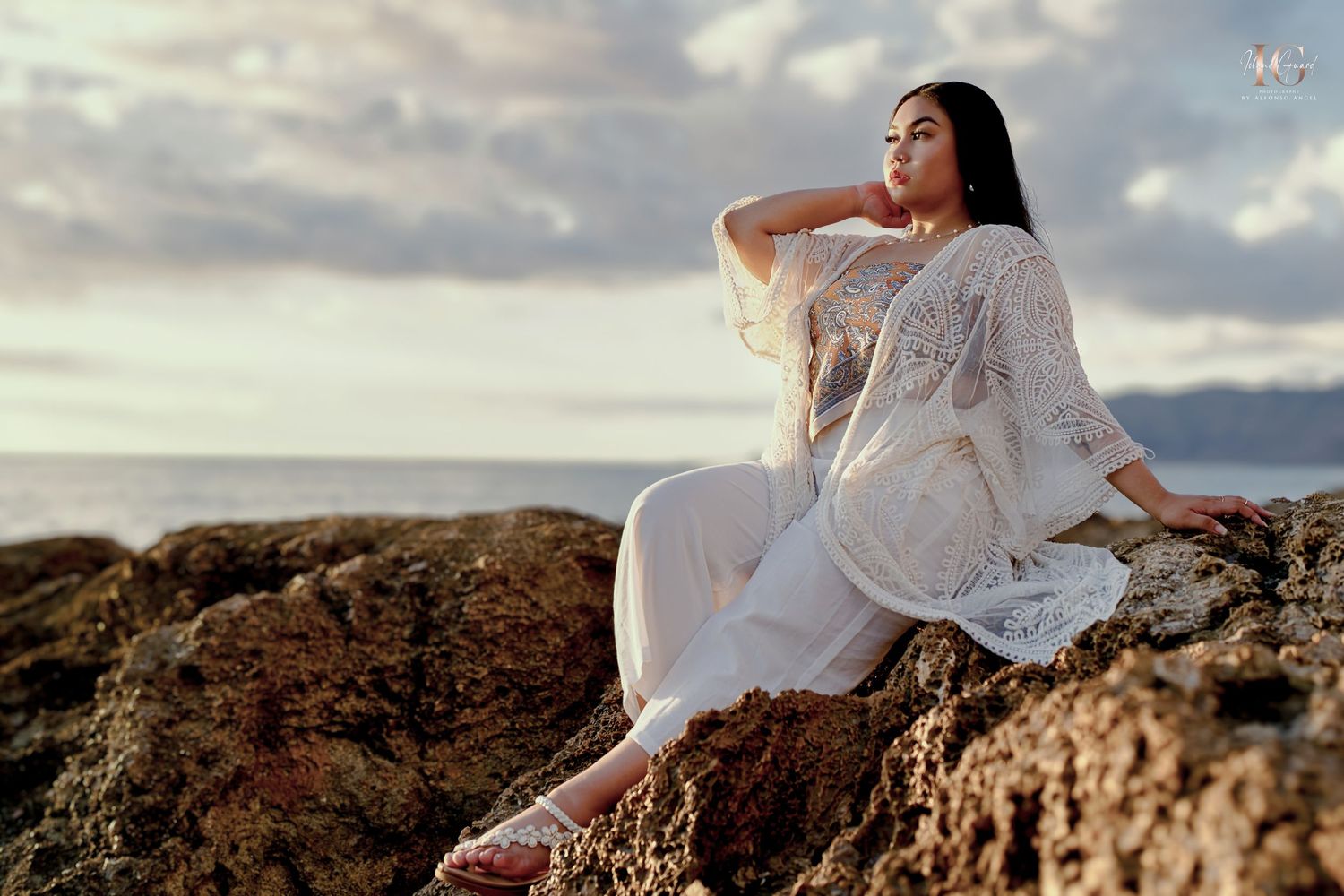 A person in a flowing white dress sits gracefully on rugged coastal rocks at sunset, looking out toward the ocean.