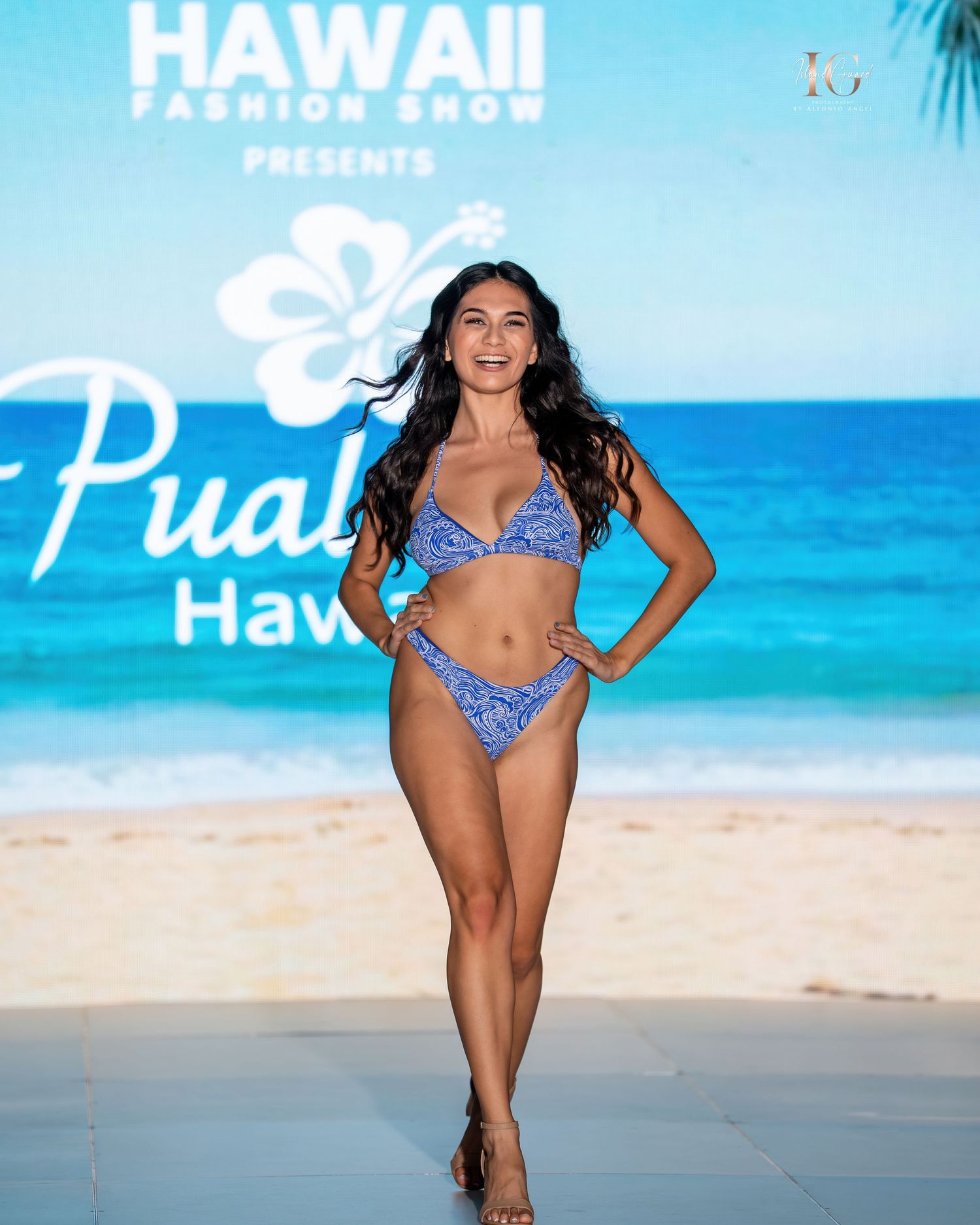 Model in blue bikini poses on scenic Hawaiian beach with turquoise ocean and palm trees in background.