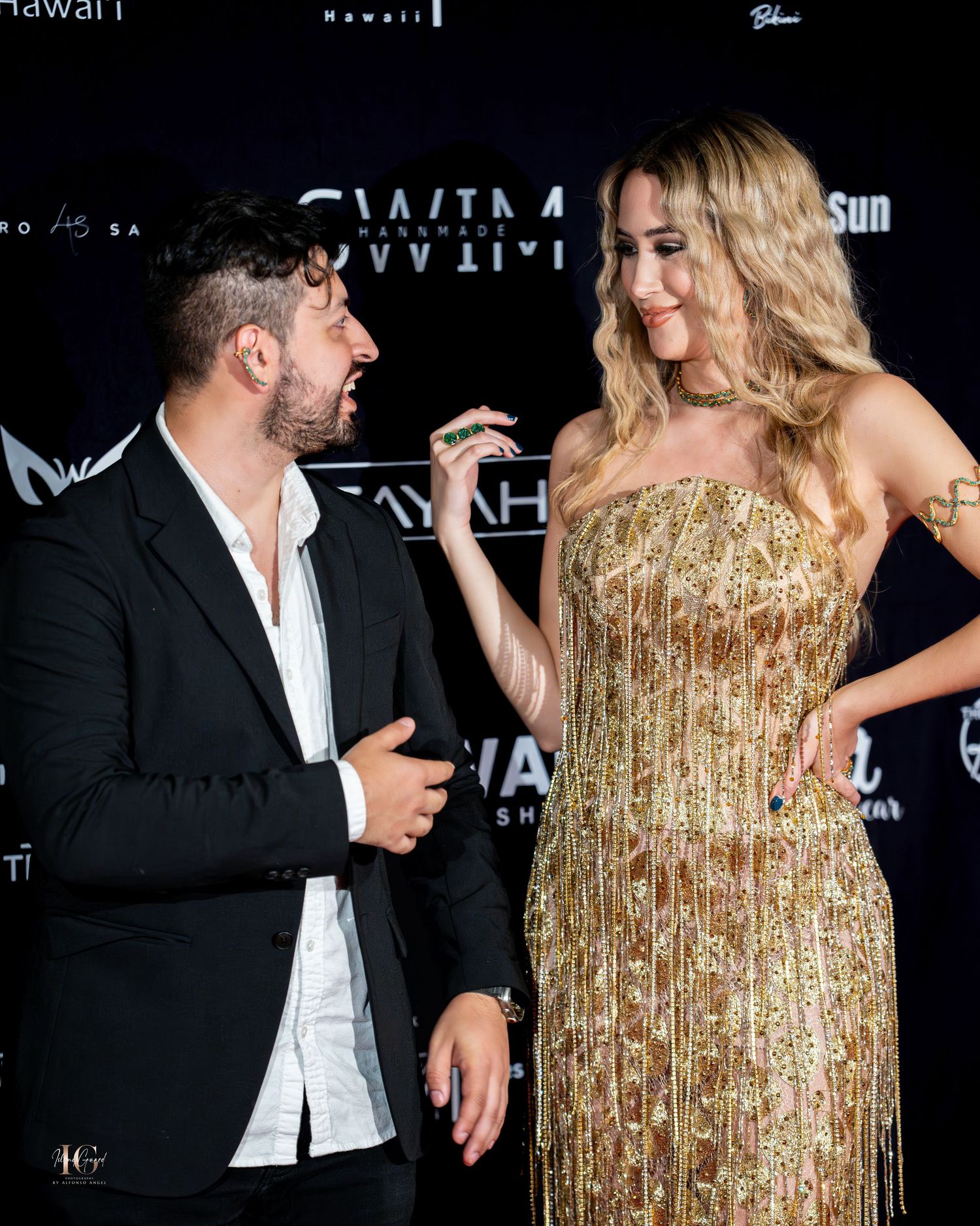 Two people in formal attire share a lighthearted moment at an event, with one wearing a sparkling gold gown.