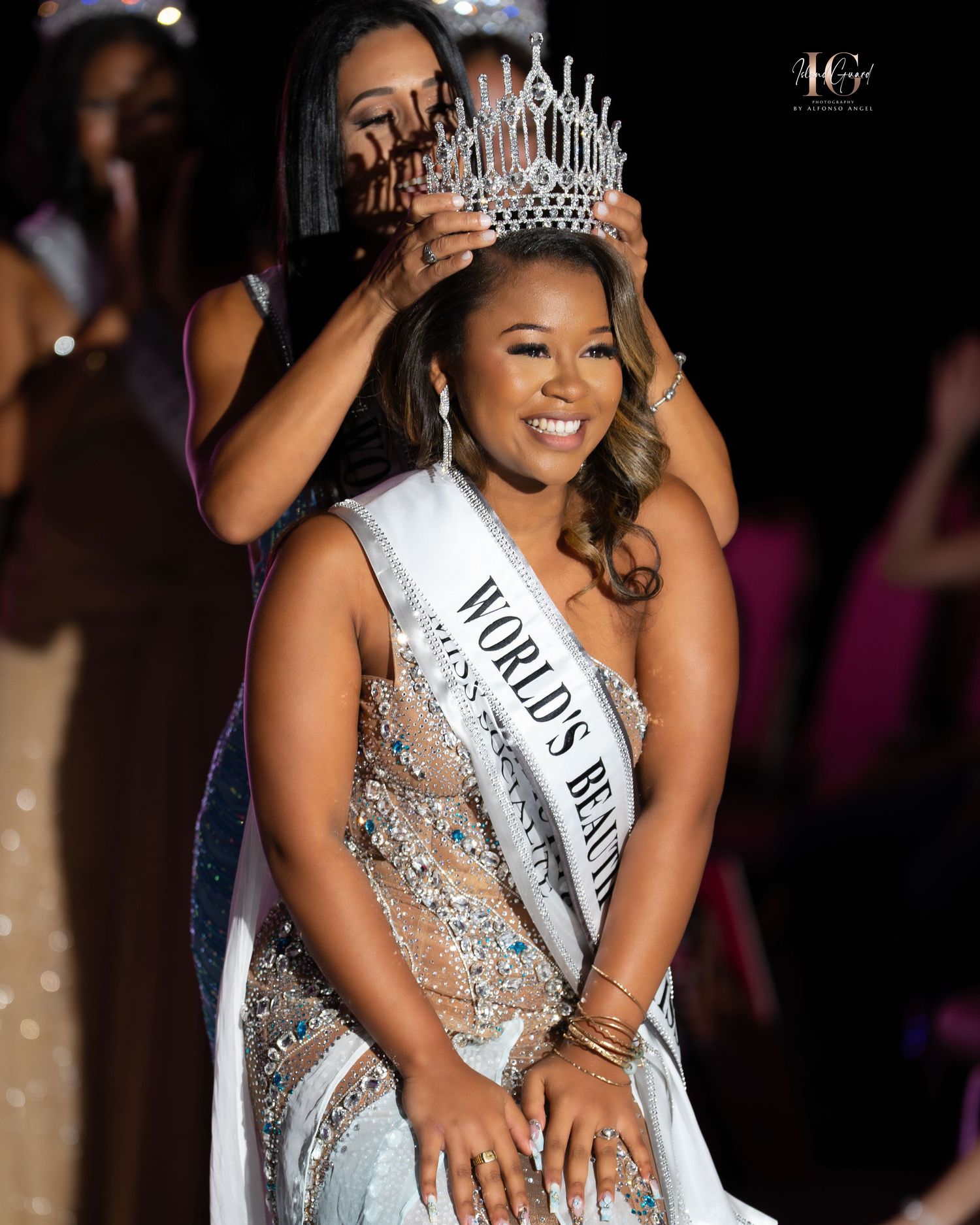 A beauty pageant contestant wearing a sparkling silver gown and crown smiles joyfully on stage during a competition.