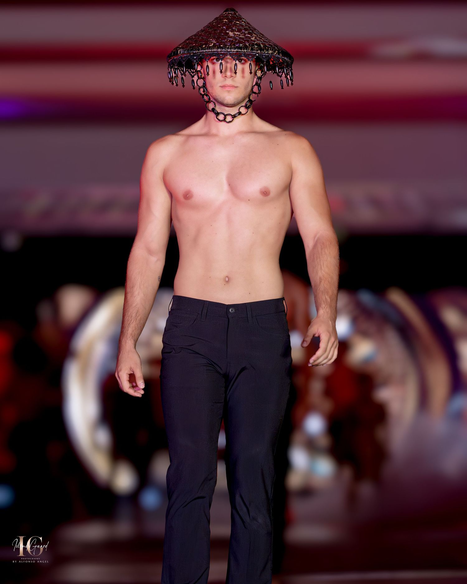 Shirtless model in dark pants and tasseled headpiece performs on fashion runway with red lighting.