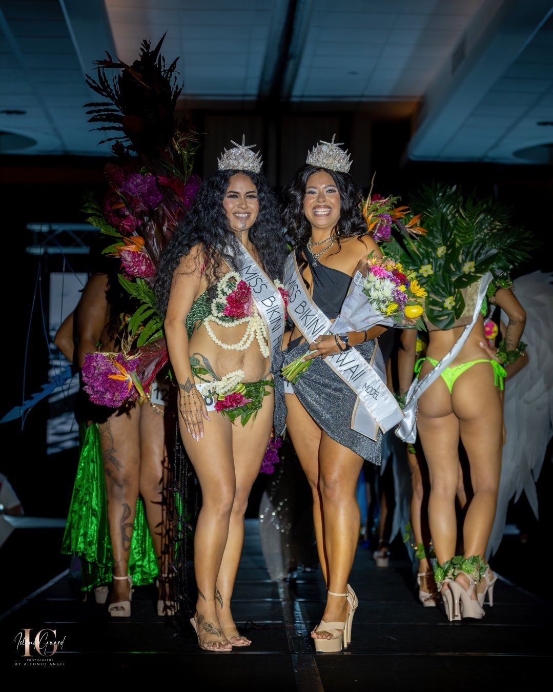 Beauty pageant winners wearing crowns and swimwear pose with colorful bouquets on stage during awards ceremony.