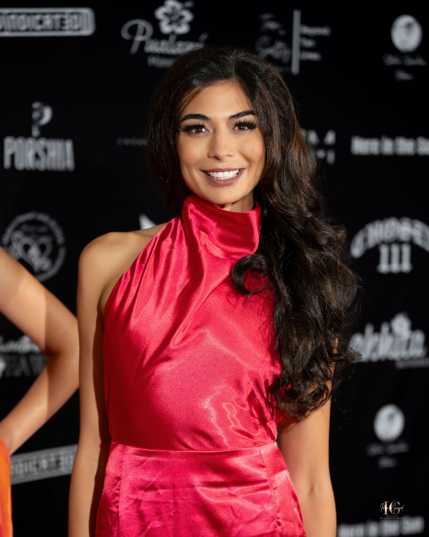 Model in red satin halter dress poses at fashion event against black backdrop.