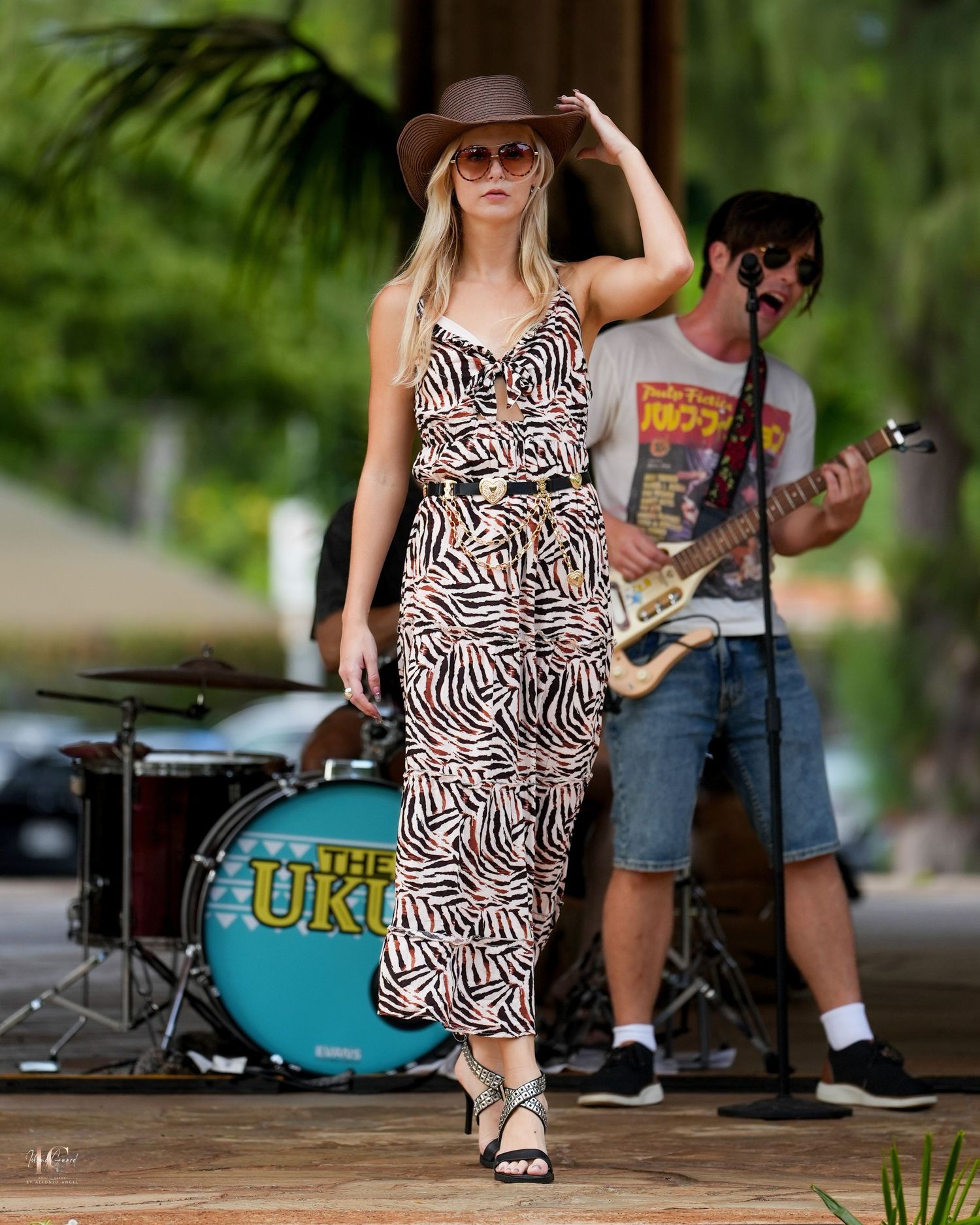 A performer in a zebra-print dress and brown hat dances on an outdoor stage during a summer concert.