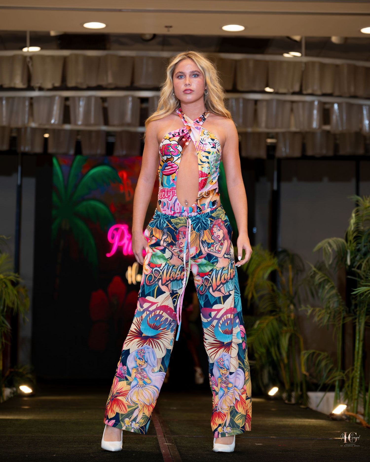 A model walks the runway in a vibrant tropical print jumpsuit at a fashion show with indoor greenery and lighting.
