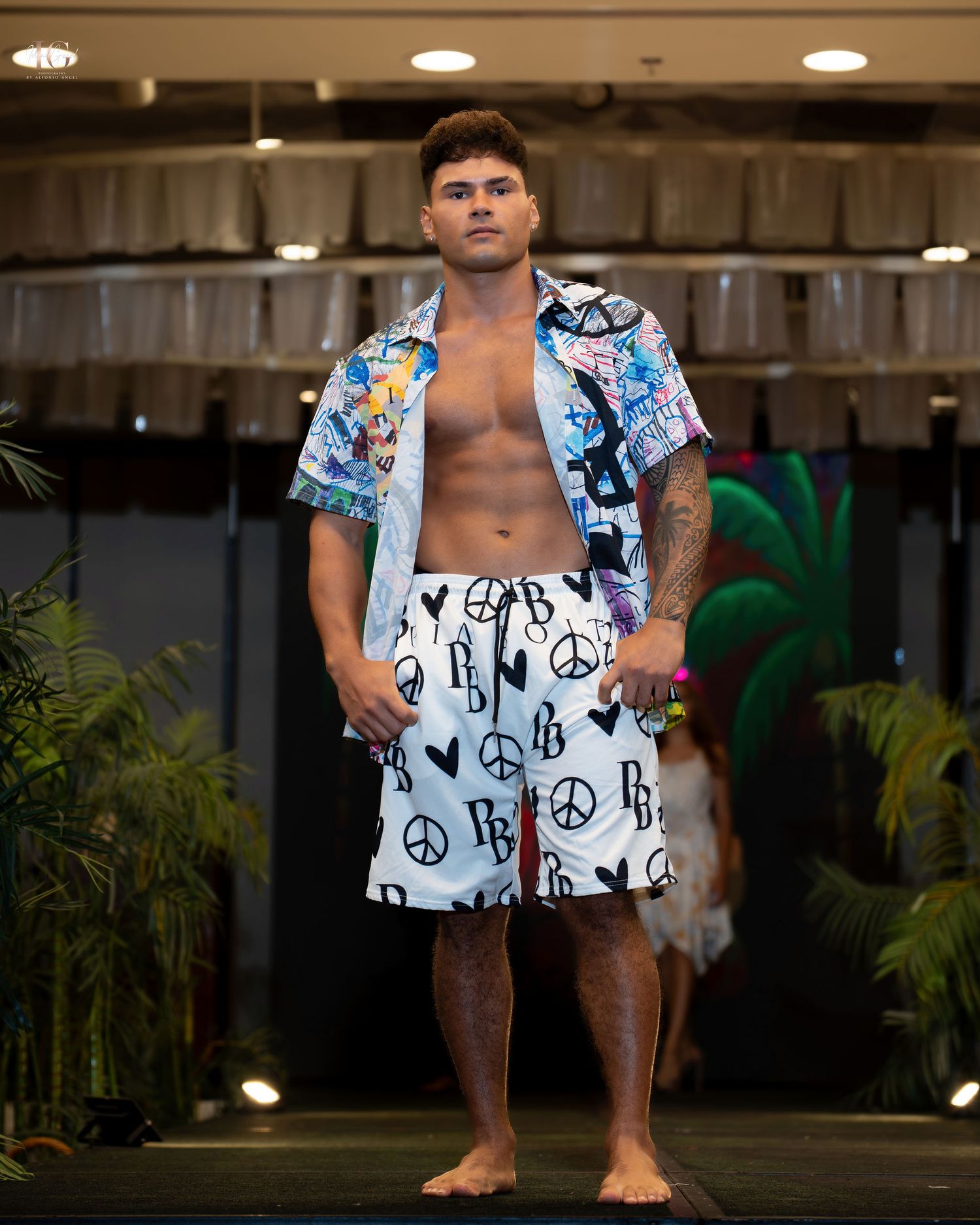 Man wearing white and black peace sign print swim shorts stands in an indoor setting with recessed lighting and plants.