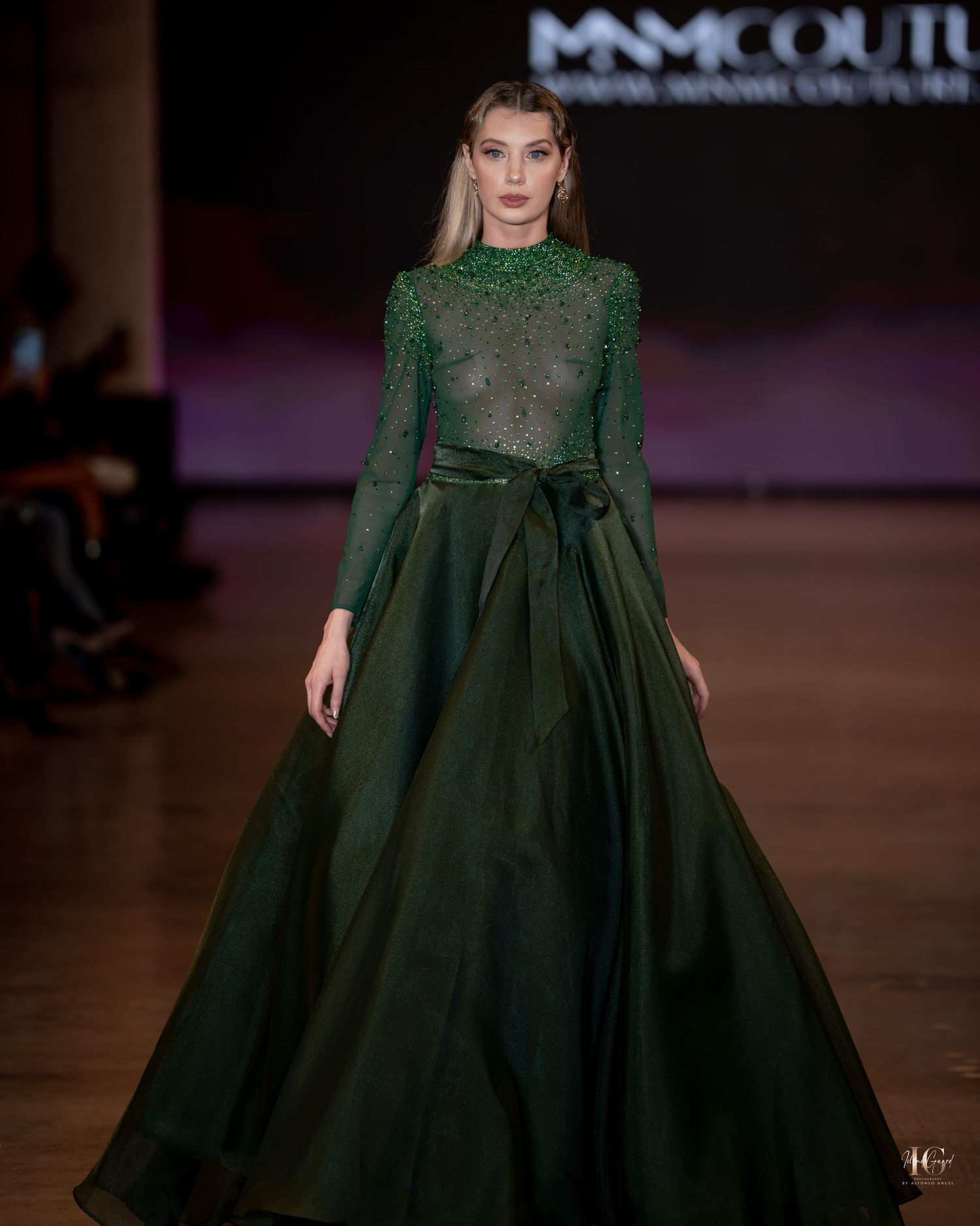 Model walks runway in elegant emerald green evening gown with long sleeves and flowing skirt during fashion show.