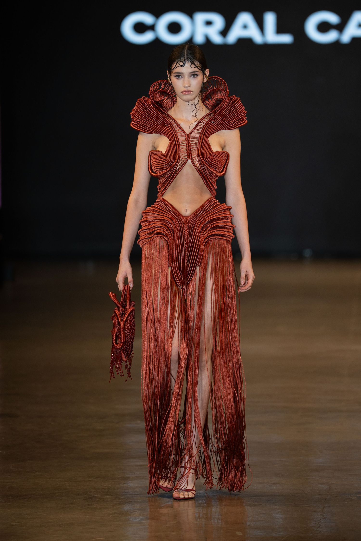 Fashion model walks runway in dramatic rust-colored fringe gown with plunging neckline at fashion show.