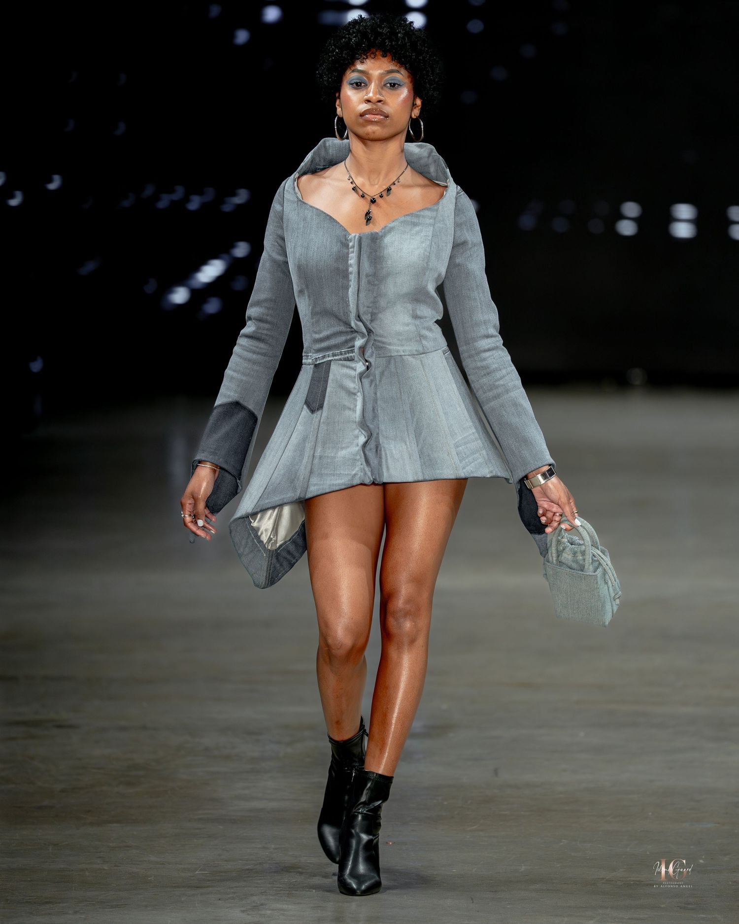 Model walks runway in gray structured blazer dress with flared sleeves and black boots at fashion show.