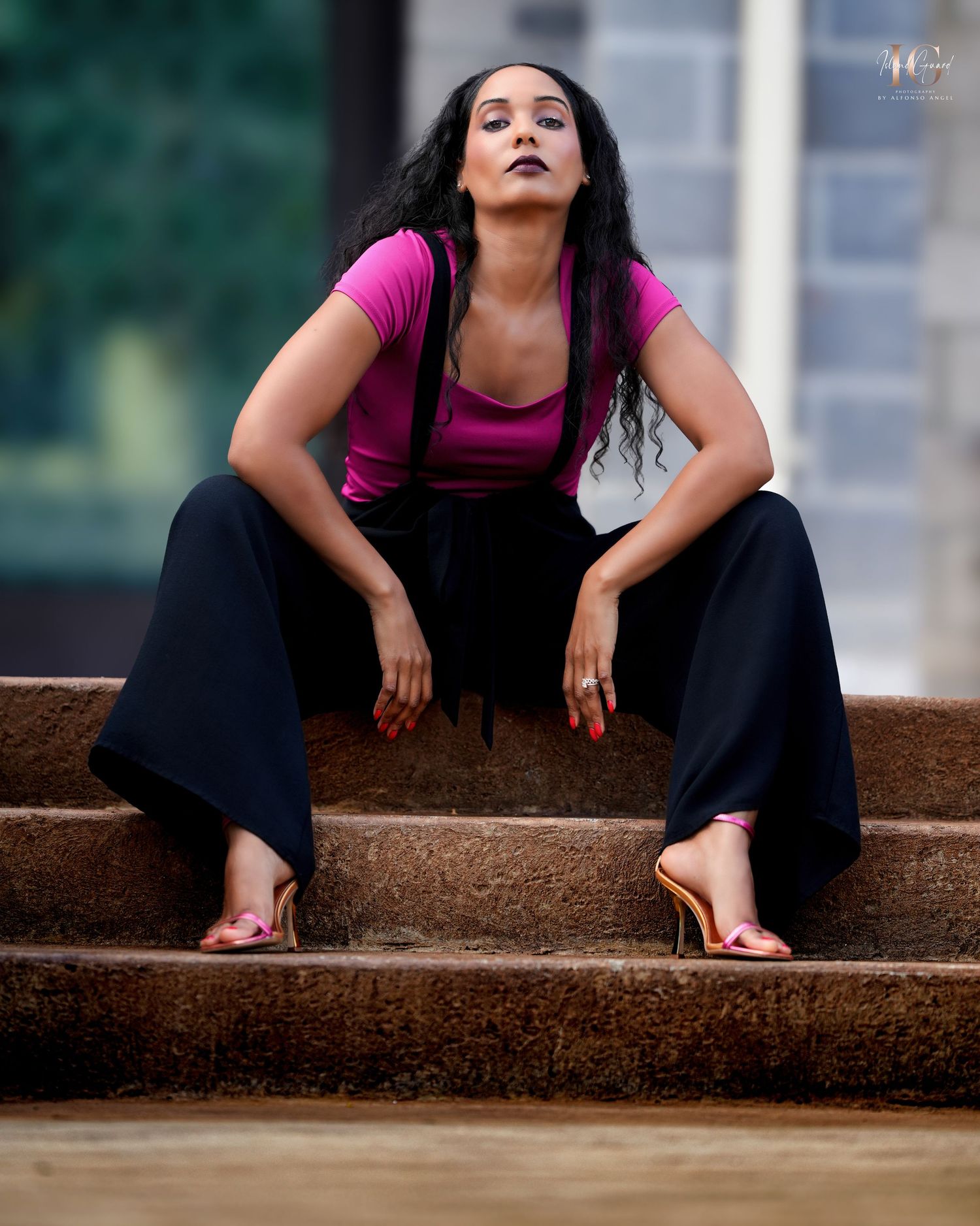 Woman in pink top and black pants sits confidently on rustic stone steps with an artistic pose.