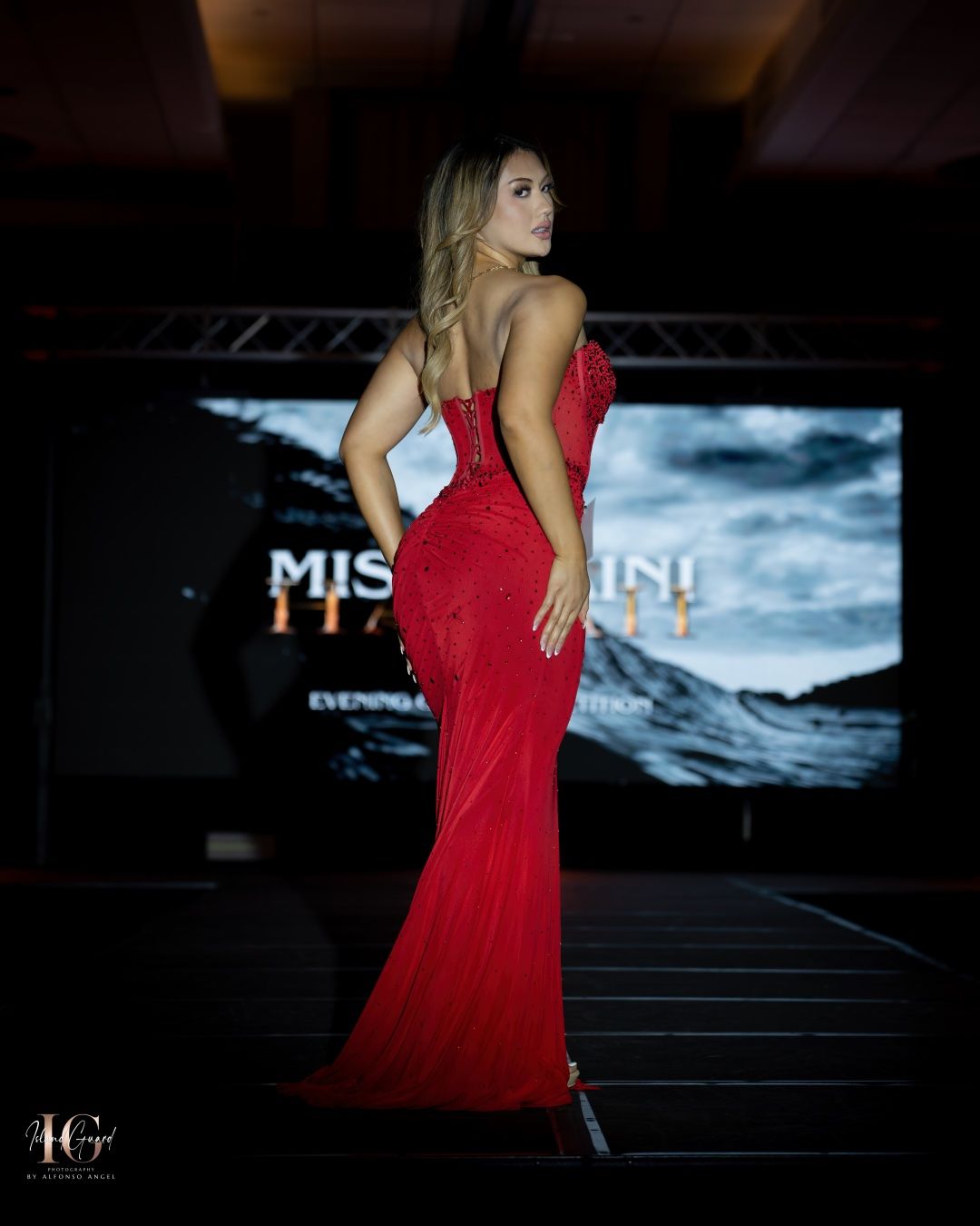 A woman poses on a runway in a form-fitting red evening gown against a dark background with illuminated signage.