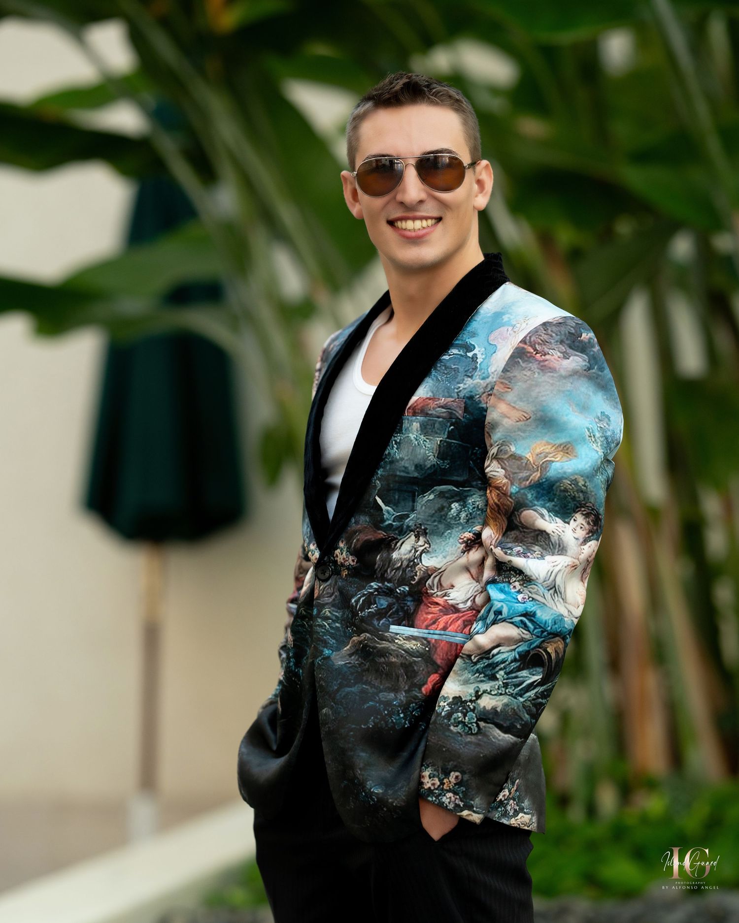 Model wearing sunglasses and printed blazer poses against tropical plant background.