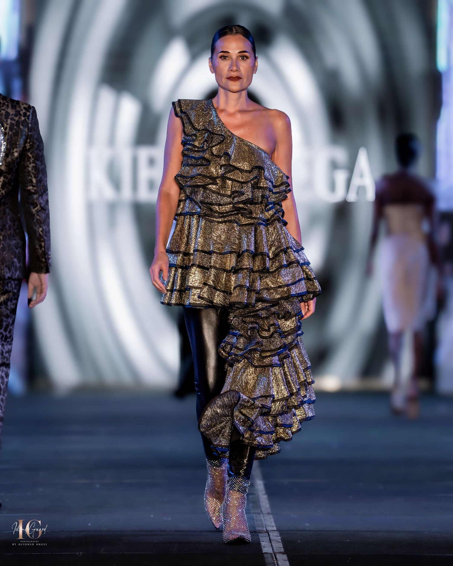Gold metallic ruffled dress shown on runway with dramatic spiral background lighting.