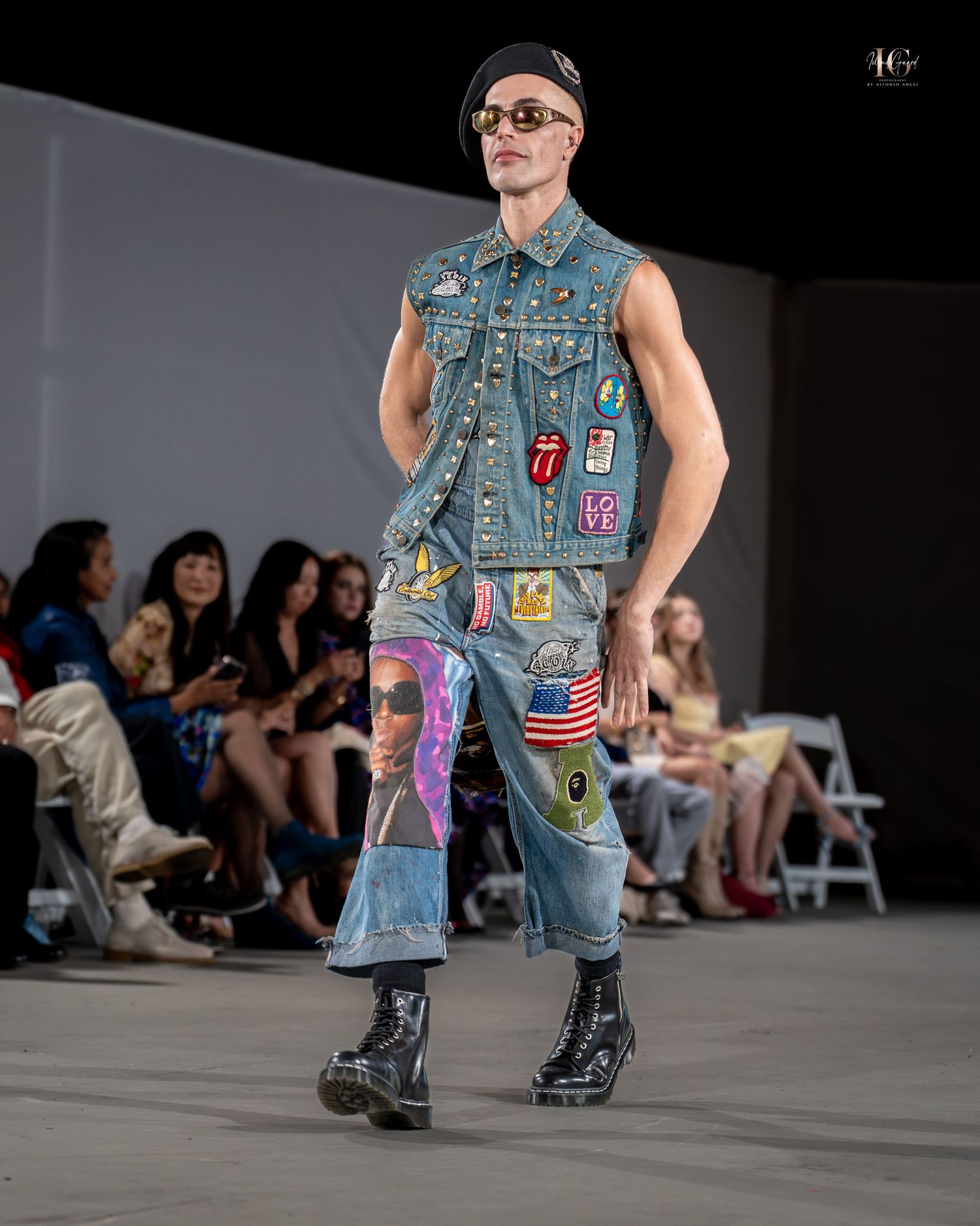 Model walks runway in distressed denim vest with colorful patches and combat boots at fashion show.
