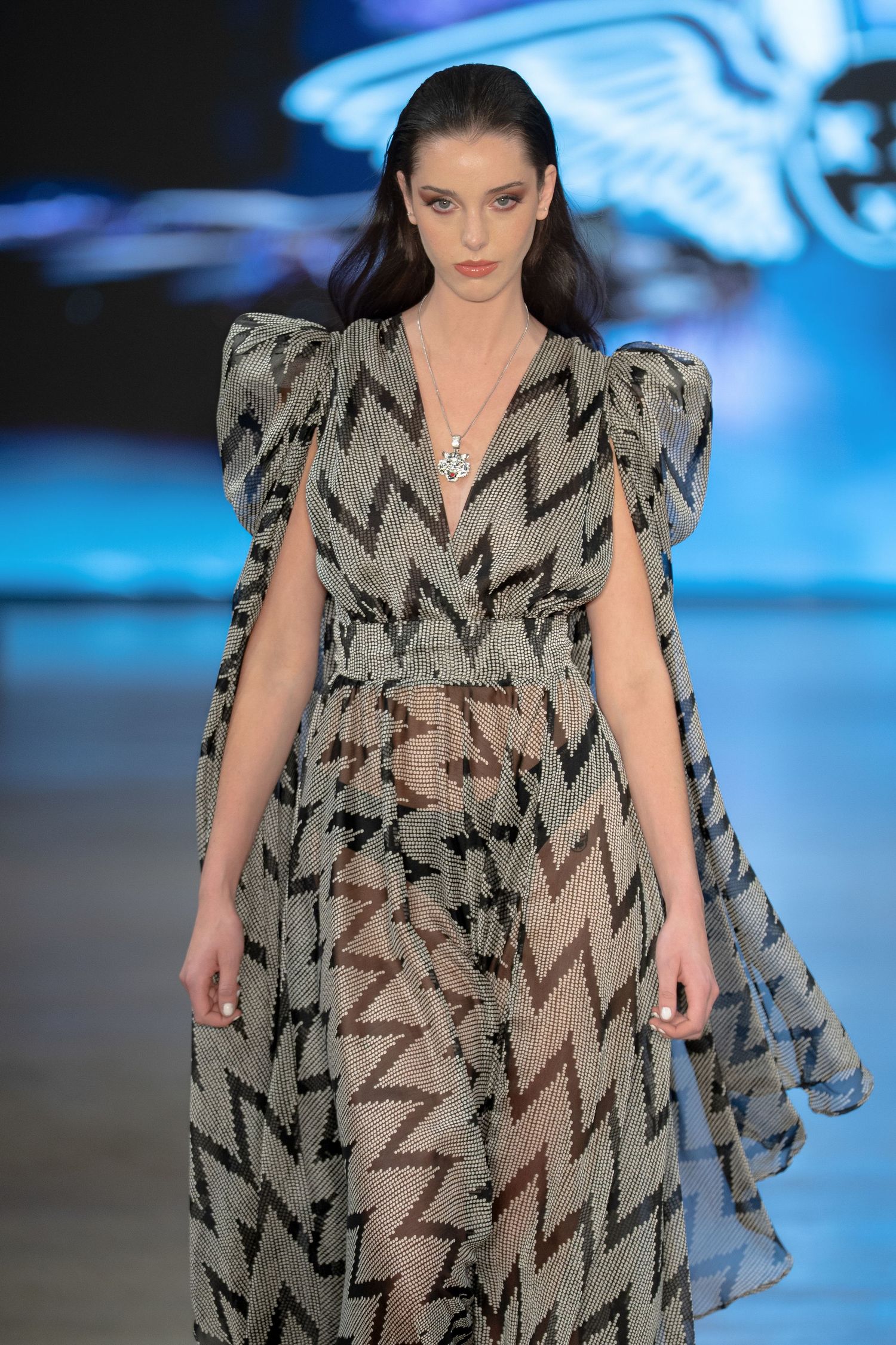 A model walks the runway in a flowing zigzag patterned dress with dramatic ruffled shoulders at a fashion show.