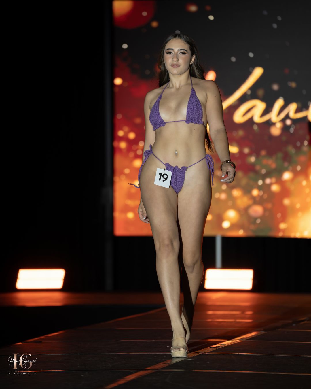Model presents purple swimsuit design on illuminated fashion runway.