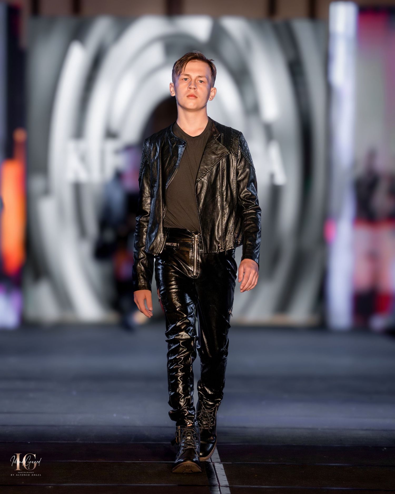 Model in black leather jacket and pants walking runway against circular backdrop lighting.