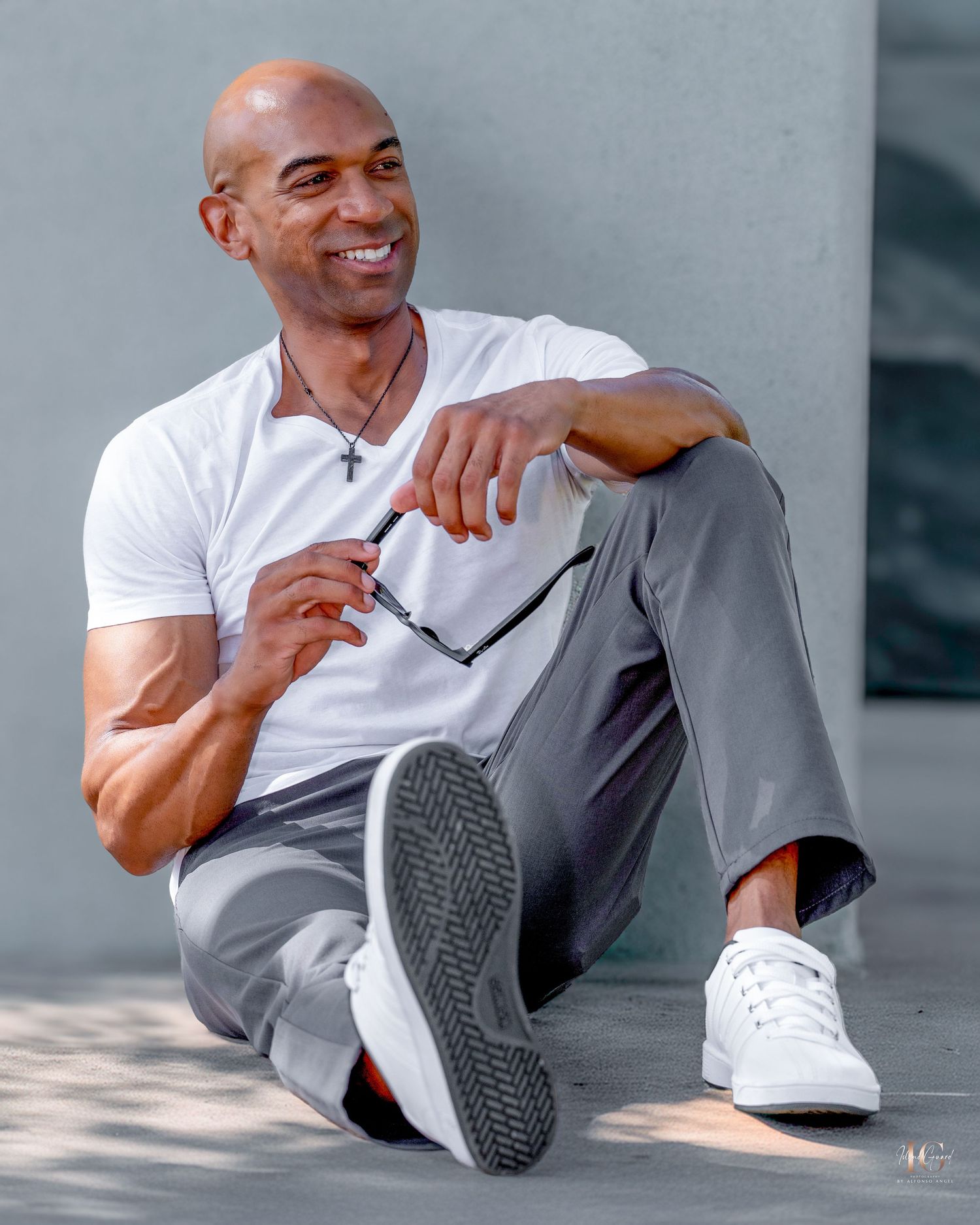 Person sitting casually against wall wearing white t-shirt and grey pants while holding glasses and smiling.