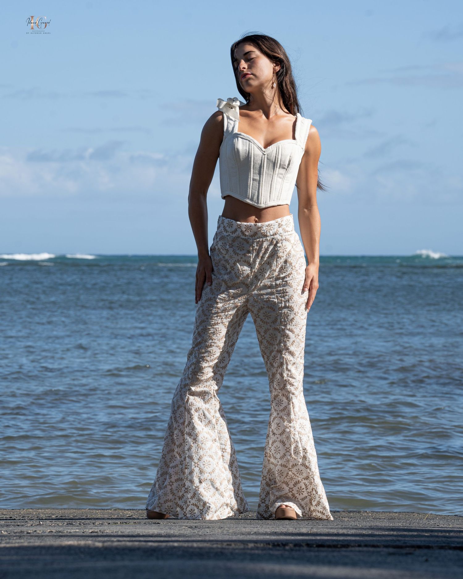 Person wearing white crop top and floral print bell bottom pants poses on a wooden dock by the ocean.