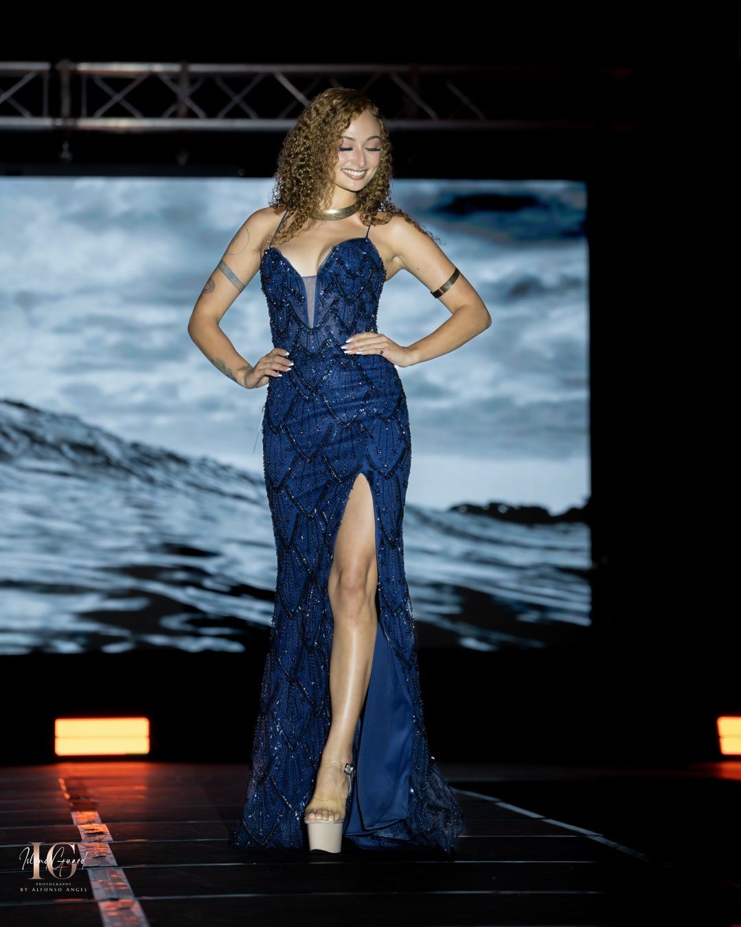 Evening gown model on runway wearing dark blue sequined dress with high slit and sweetheart neckline against ocean backdrop.