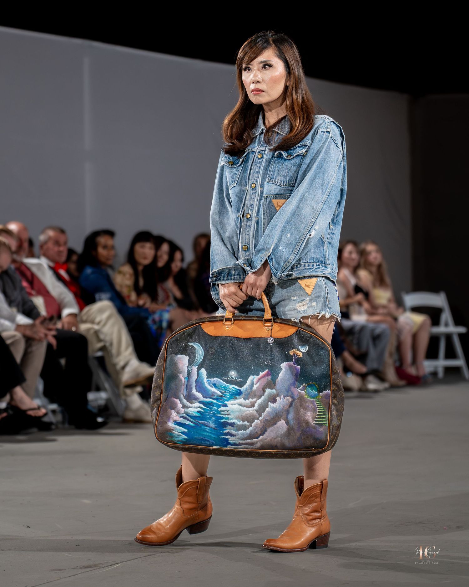Model on fashion runway wearing denim shirt and metallic skirt with brown cowboy boots and large handbag.