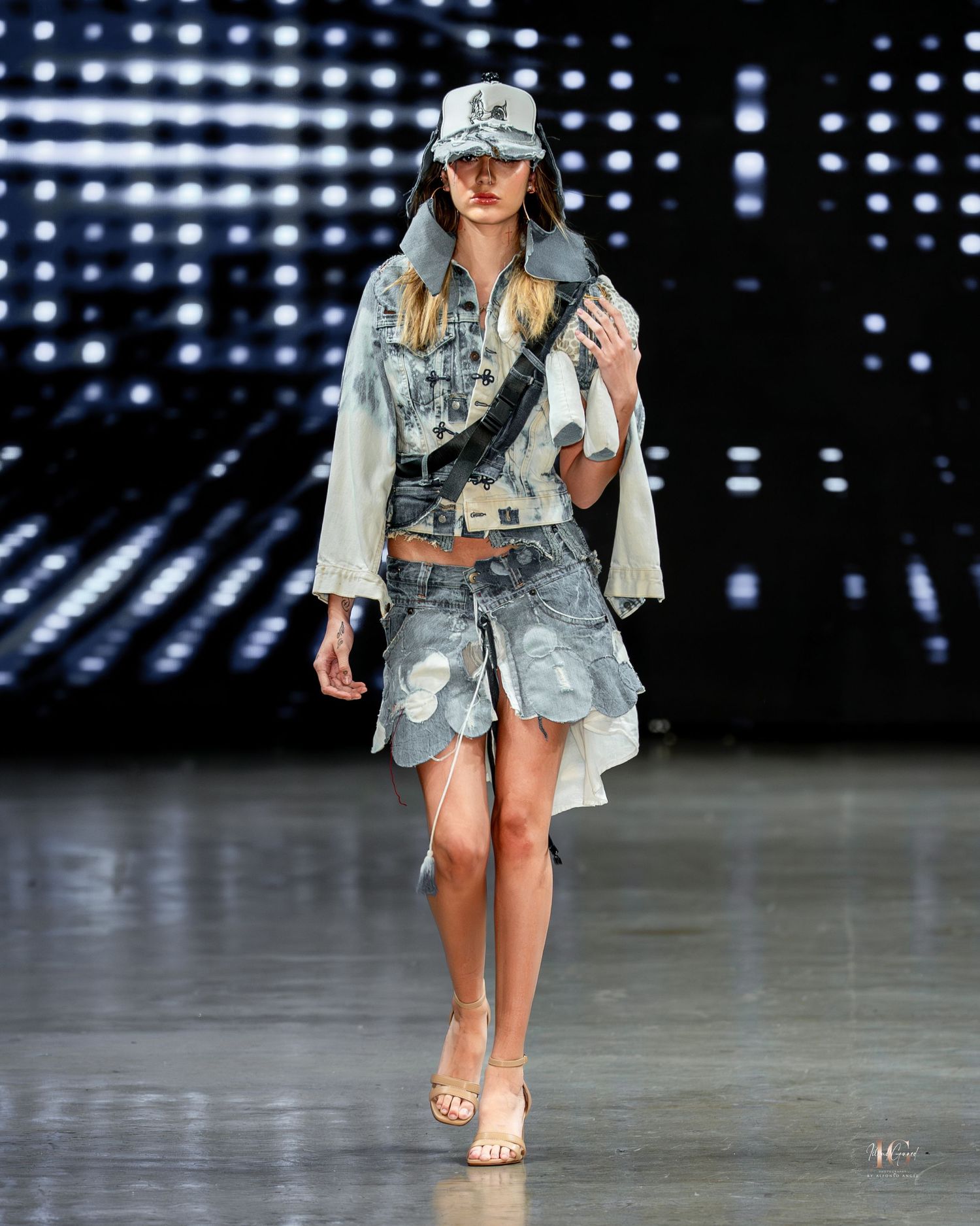Fashion model walks runway in distressed denim and white jacket ensemble during fashion show with illuminated backdrop.