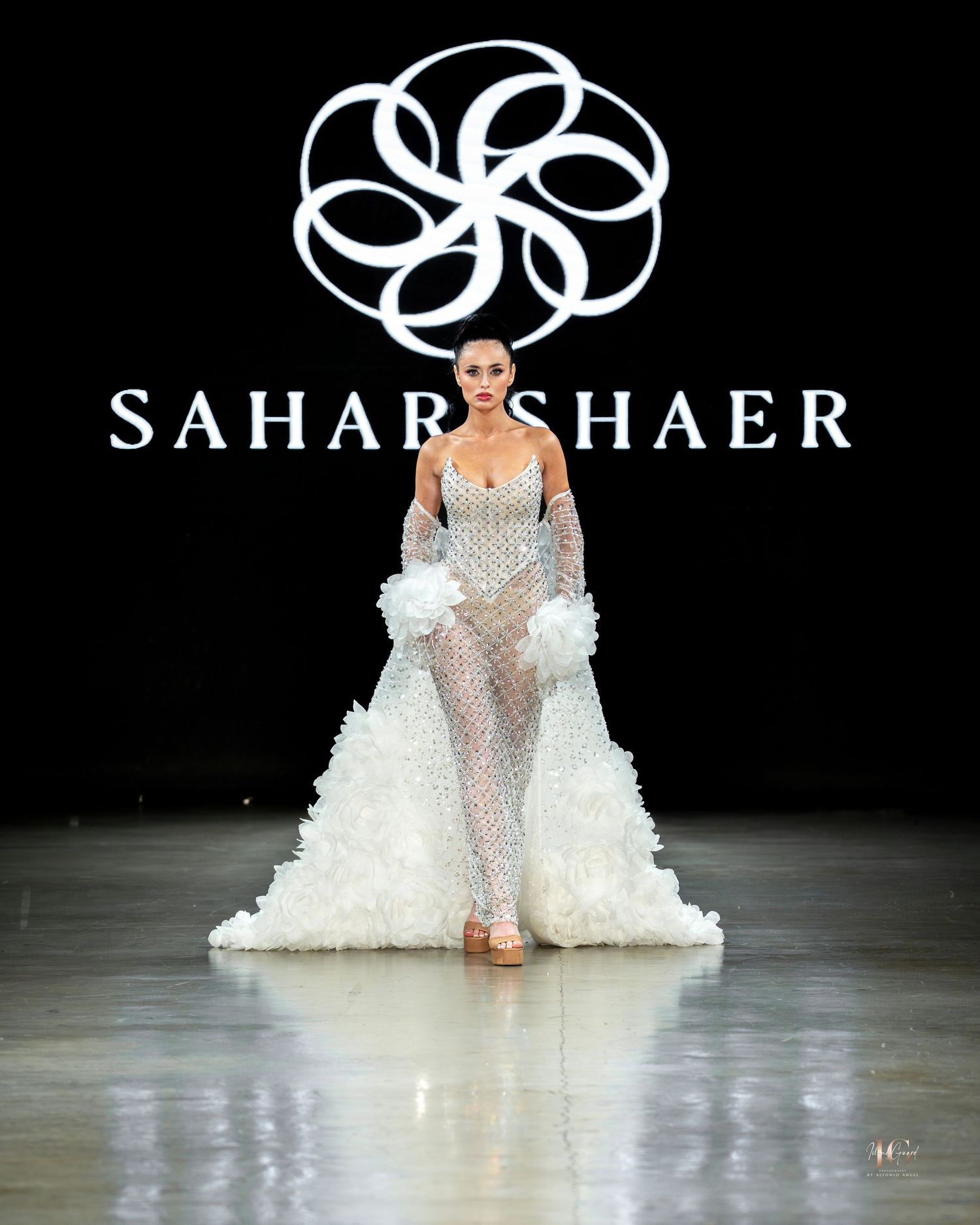 A fashion runway showcasing a flowing white bridal gown with sheer details and a dramatic train on a dark stage.