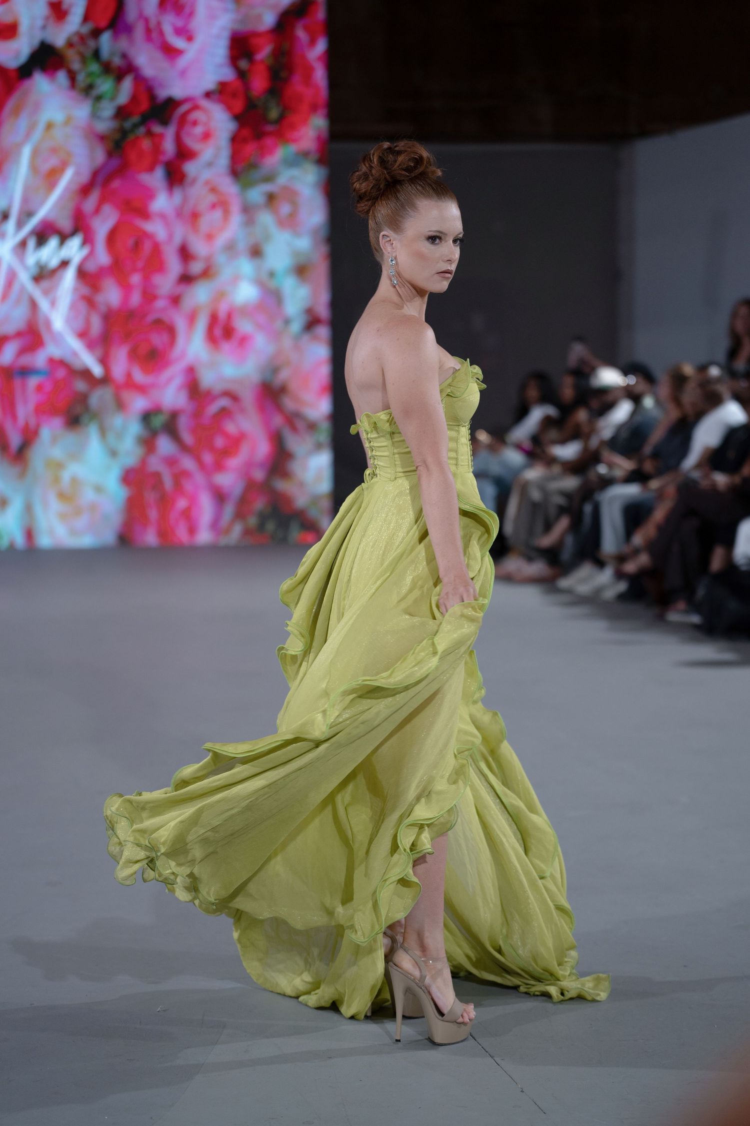 Model walks runway in flowing chartreuse evening gown with dramatic movement during fashion show.