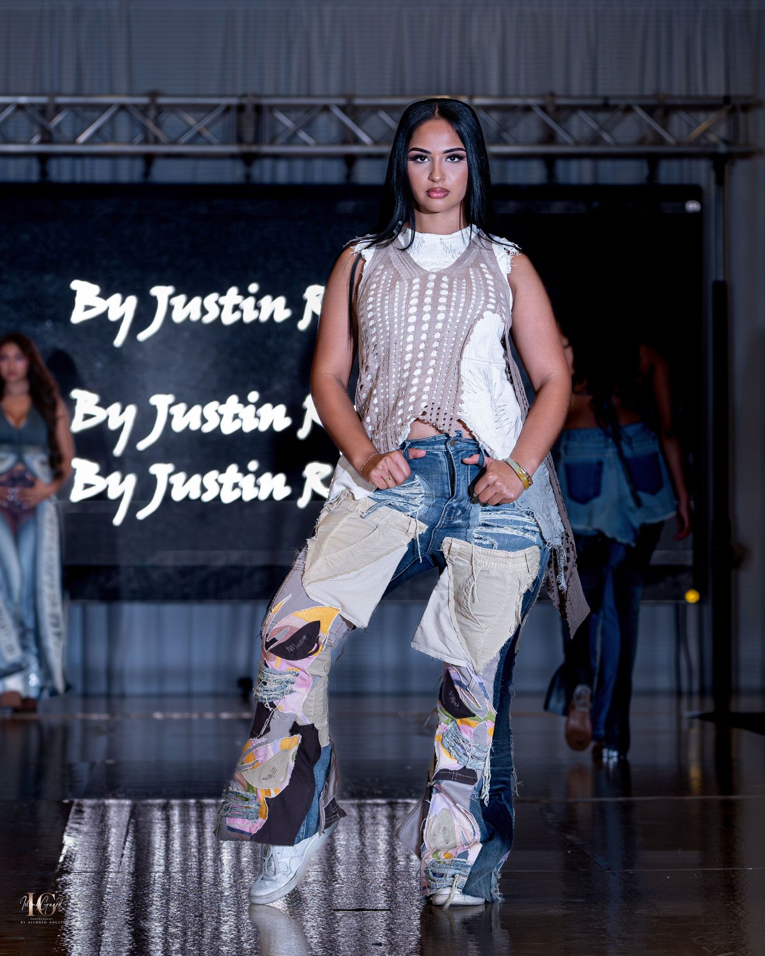 Models showcase casual streetwear outfits with denim and knitwear on a dimly lit runway during fashion show.