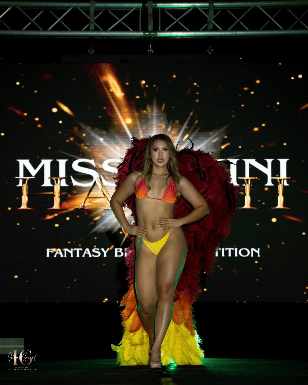 A performer in red and yellow costume with feathered accents poses on stage with dramatic lighting effects and signage.