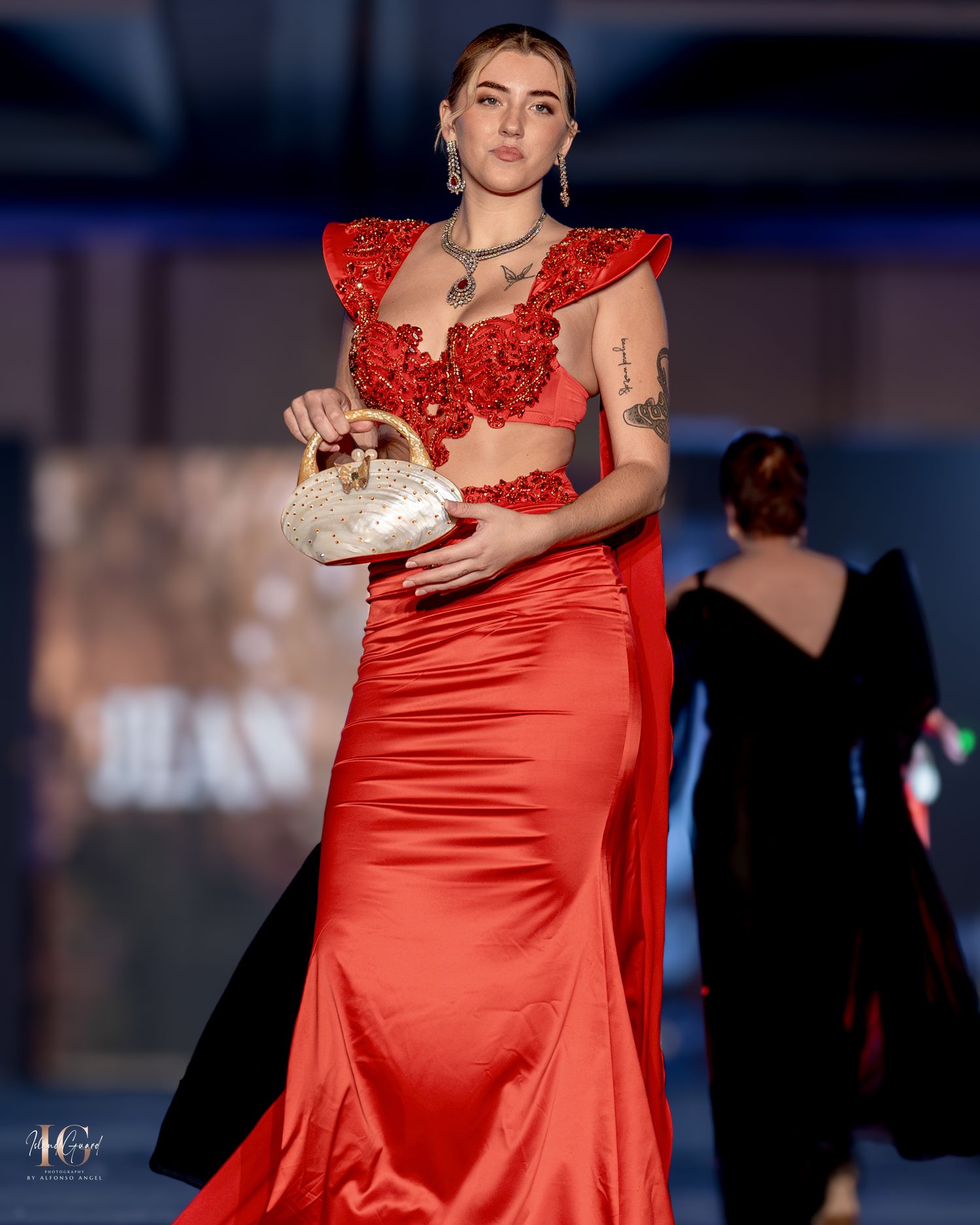 Models present flowing red satin evening gowns on modern fashion runway with theatrical lighting.