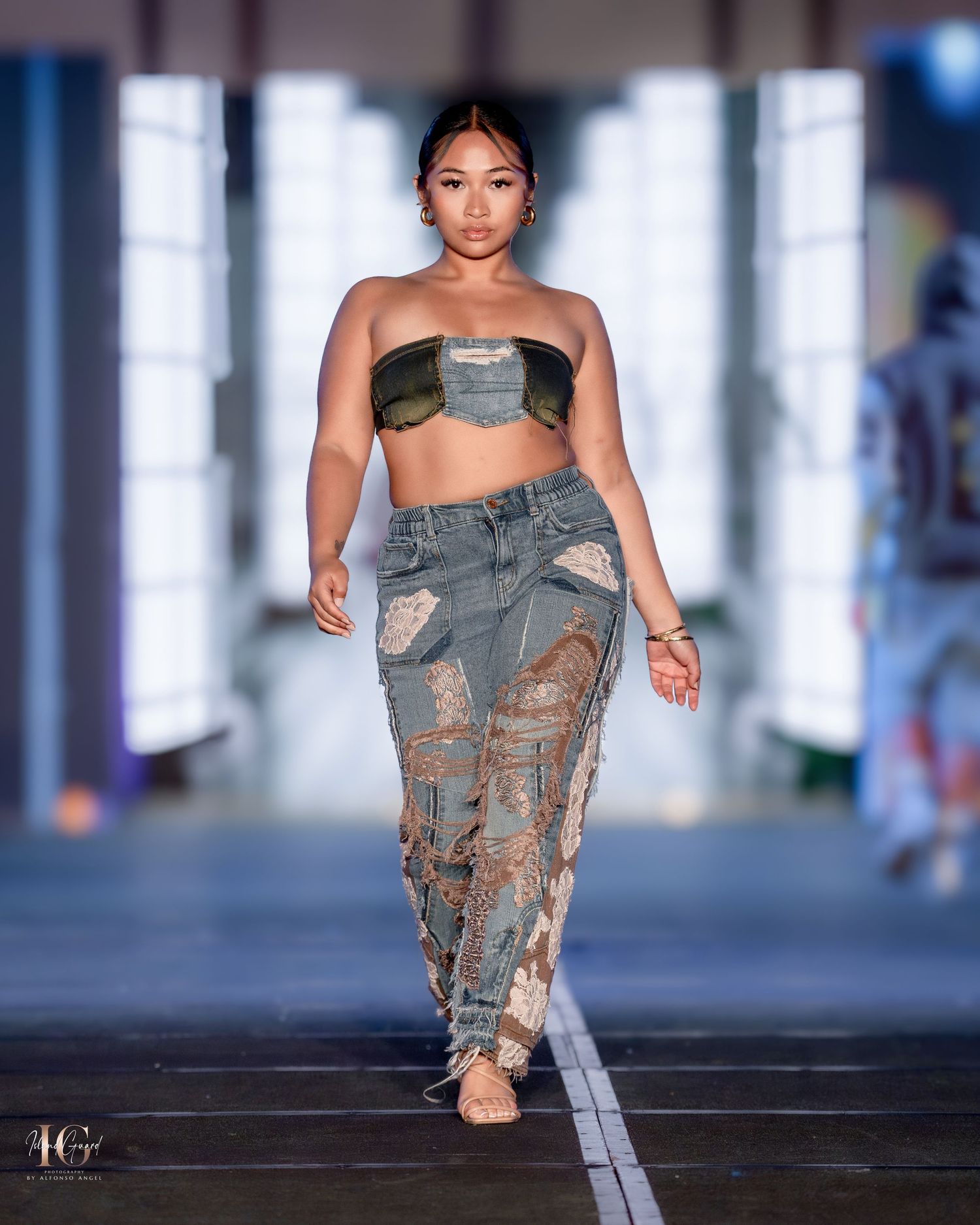 Model presents deconstructed denim look with dramatic distressing on illuminated fashion runway.