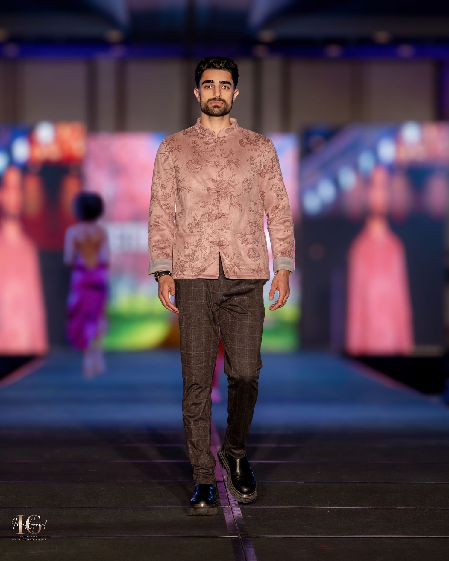 Model showcases dusty pink textured jacket with dark trousers on fashion runway.