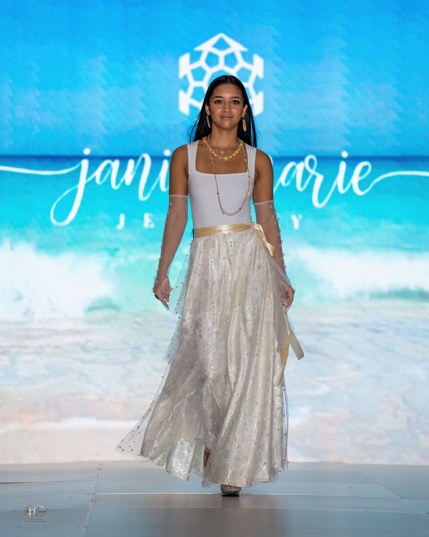 Model in white crop top and flowing metallic skirt walks runway at beachy fashion show.