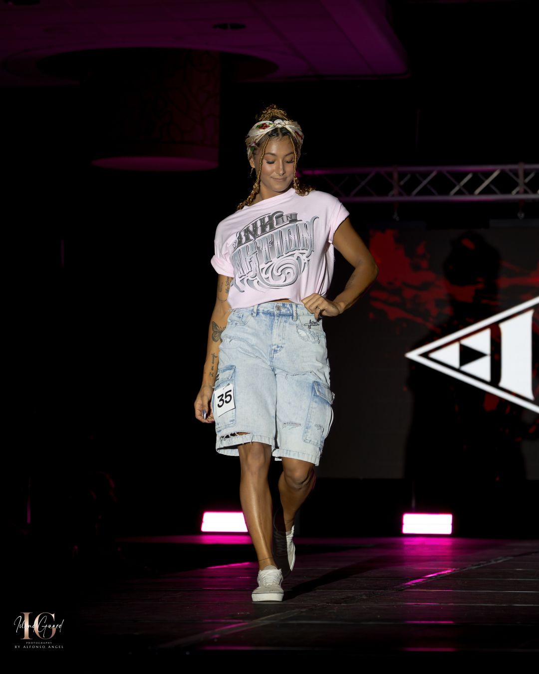 Model presents distressed denim shorts and white graphic tee on illuminated catwalk.